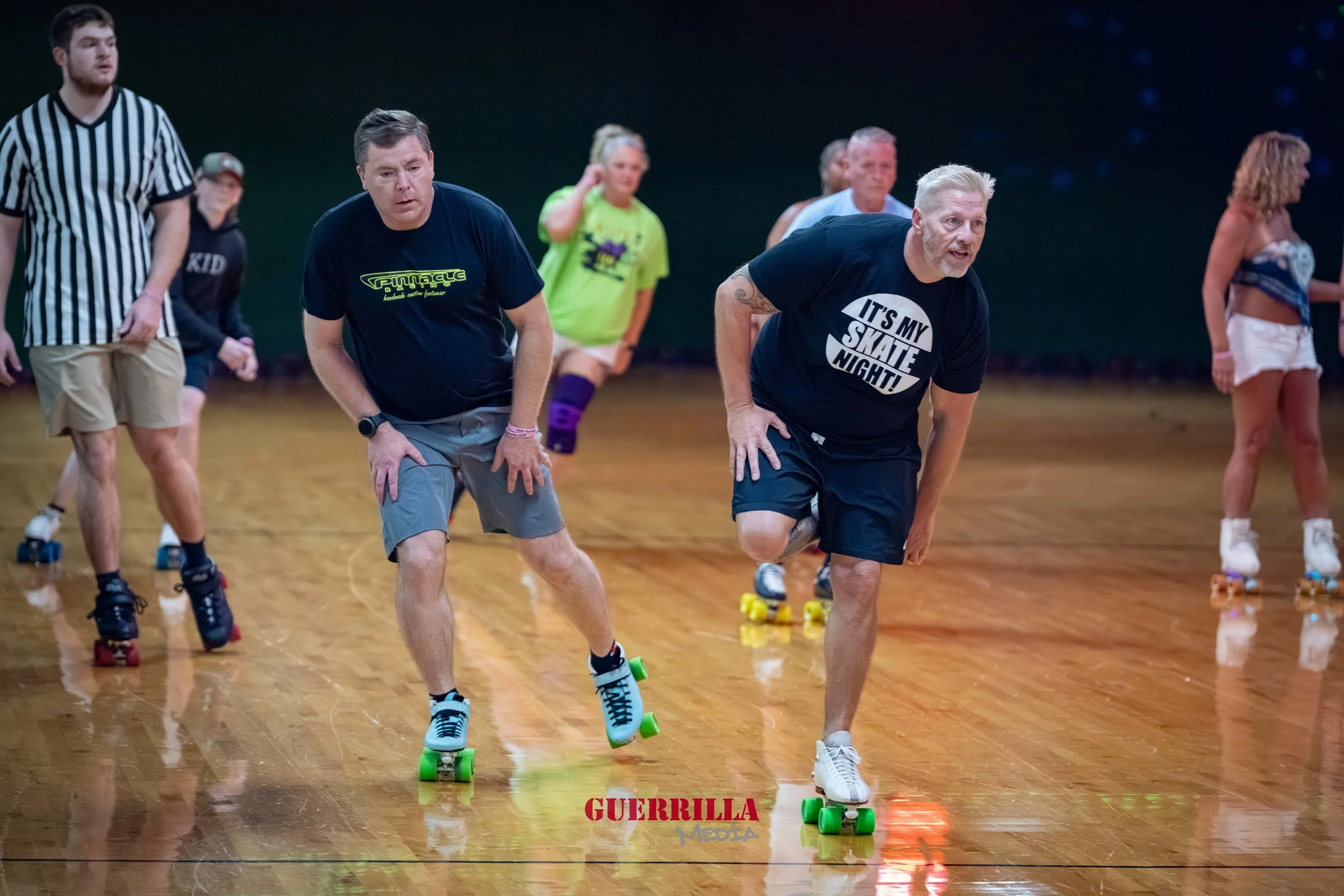 People roller skating on an indoor rink, with two men in the foreground leaning forward on their skates, one wearing a black T-shirt with the text 'It's my skate night!' and the other a navy T-shirt, with several women and a referee in the background