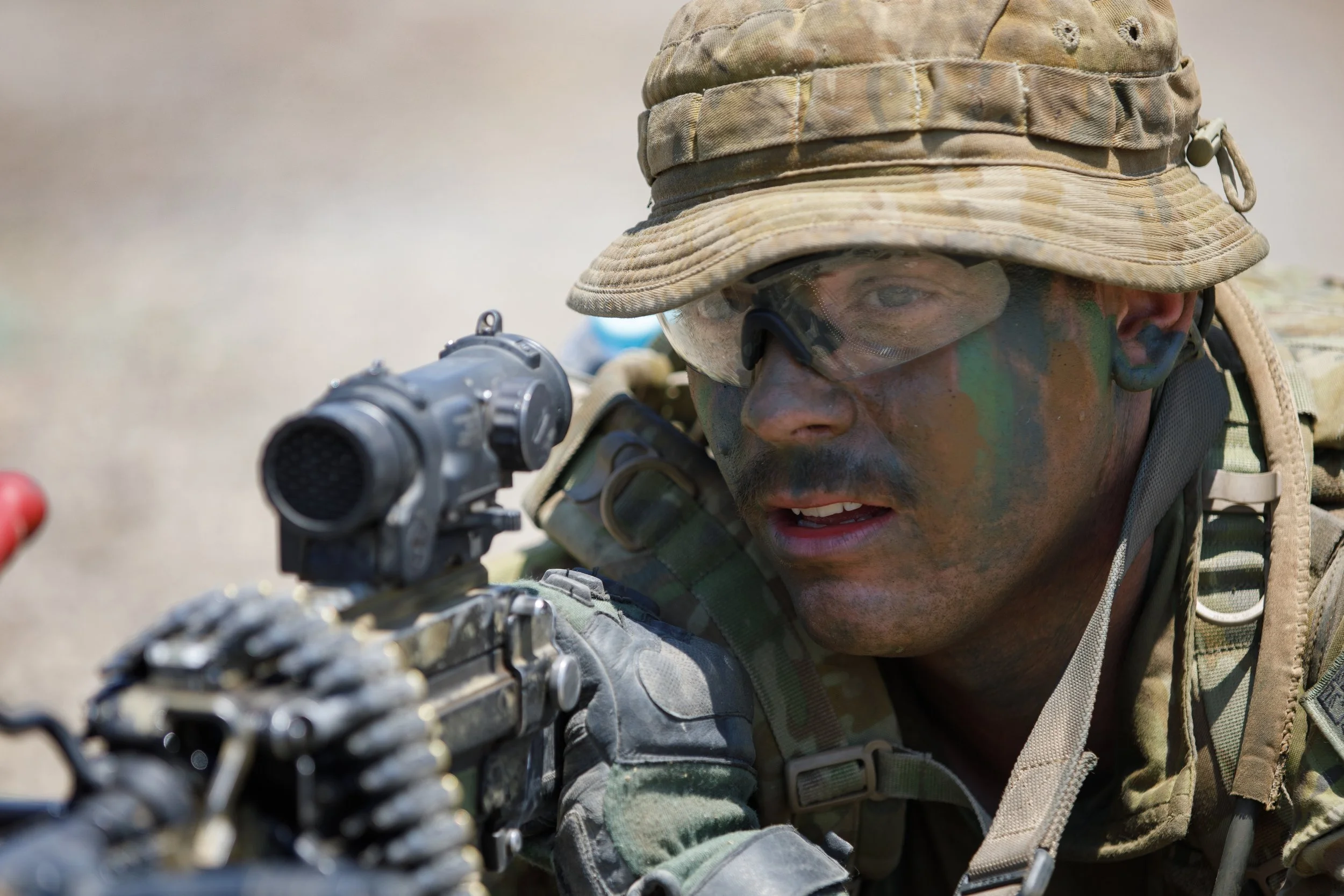 A soldier wearing camouflage face paint, glasses, and military gear, aiming with a gun.