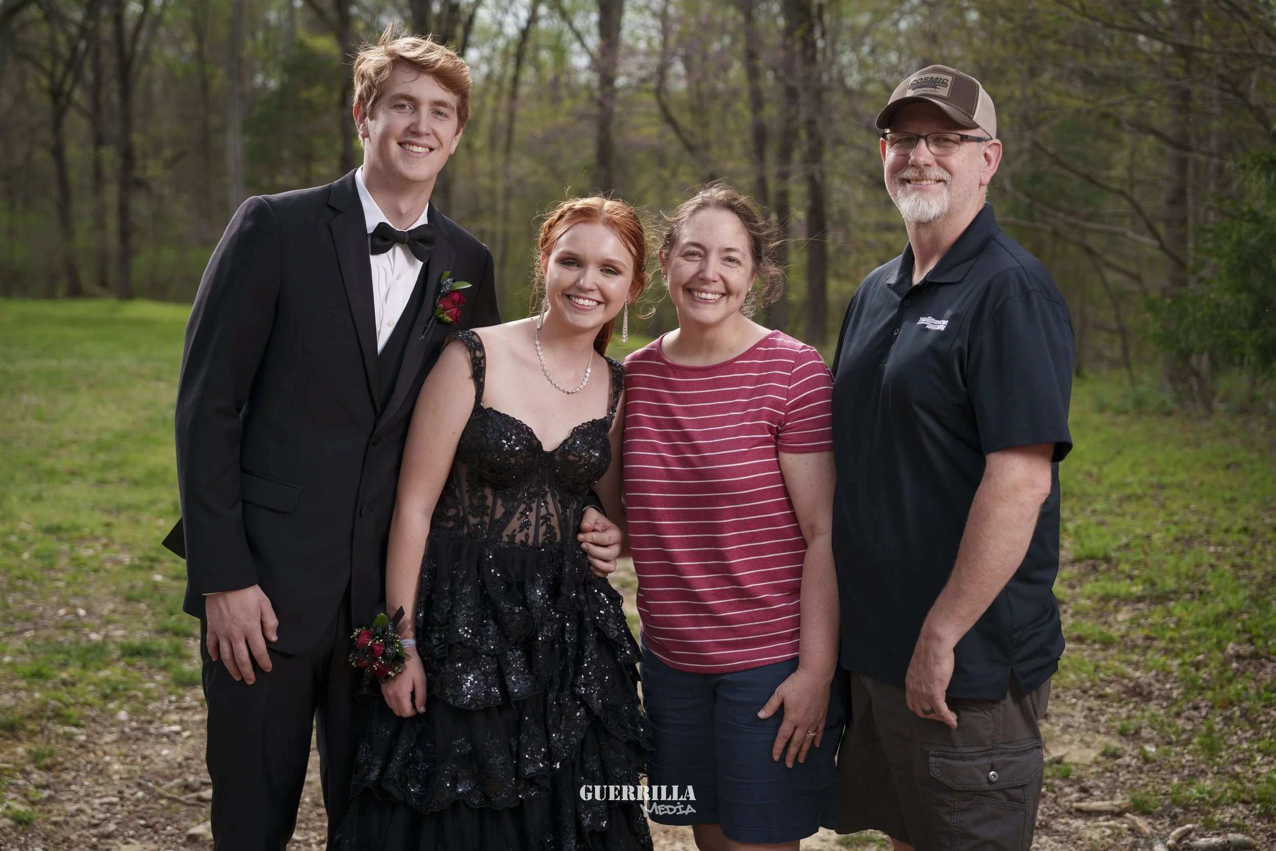 A group photo of four people outdoors in a wooded area, smiling, with a young man in a tuxedo and a young woman in a black dress, alongside two adults, a woman in a red striped shirt and a man in a black shirt and cap.
