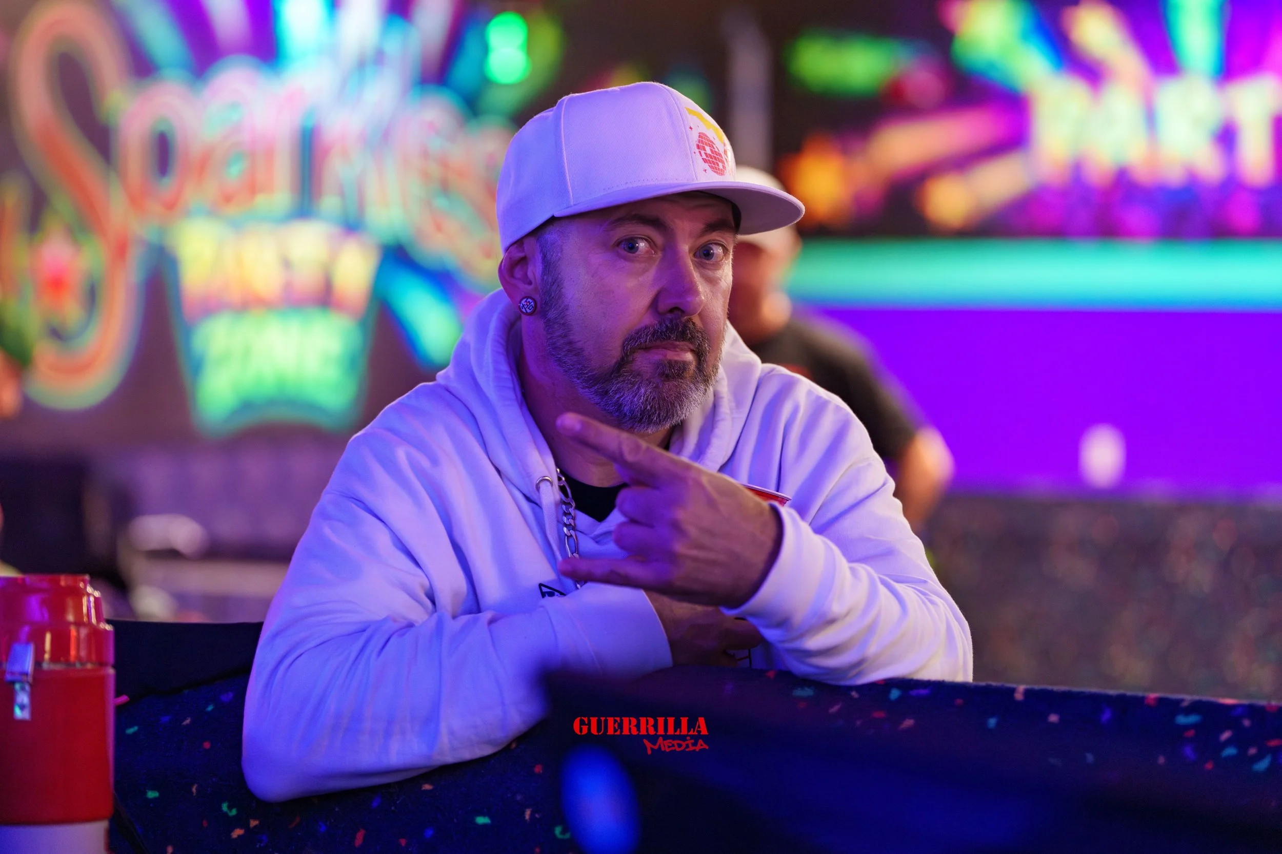 A middle-aged man with a beard and earrings, wearing a white hoodie and a white cap, sits at a table in a neon-lit arcade. He is pointing at himself or making a gesture with his hand, with a serious expression. The background features colorful, blurr