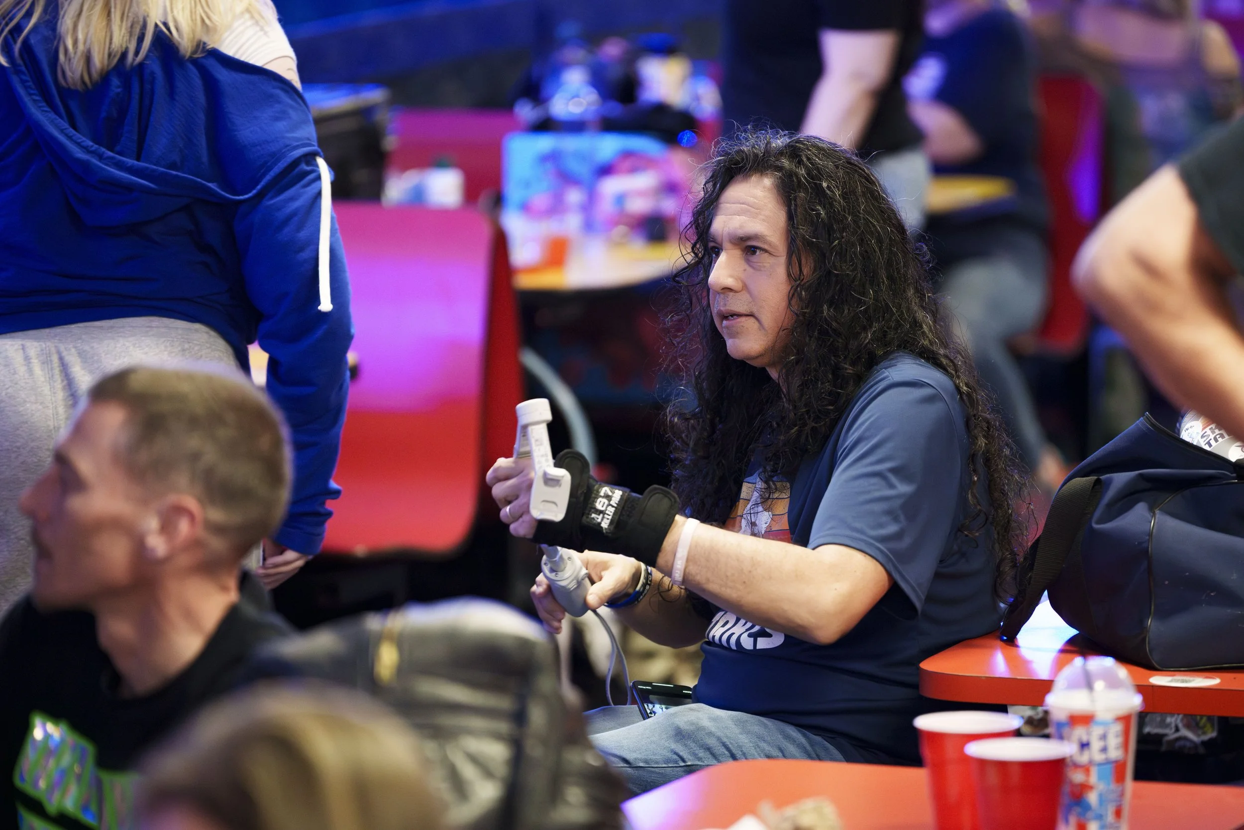 A person with long hair sitting at a table, holding an electrical device, surrounded by others at a social event or gathering.