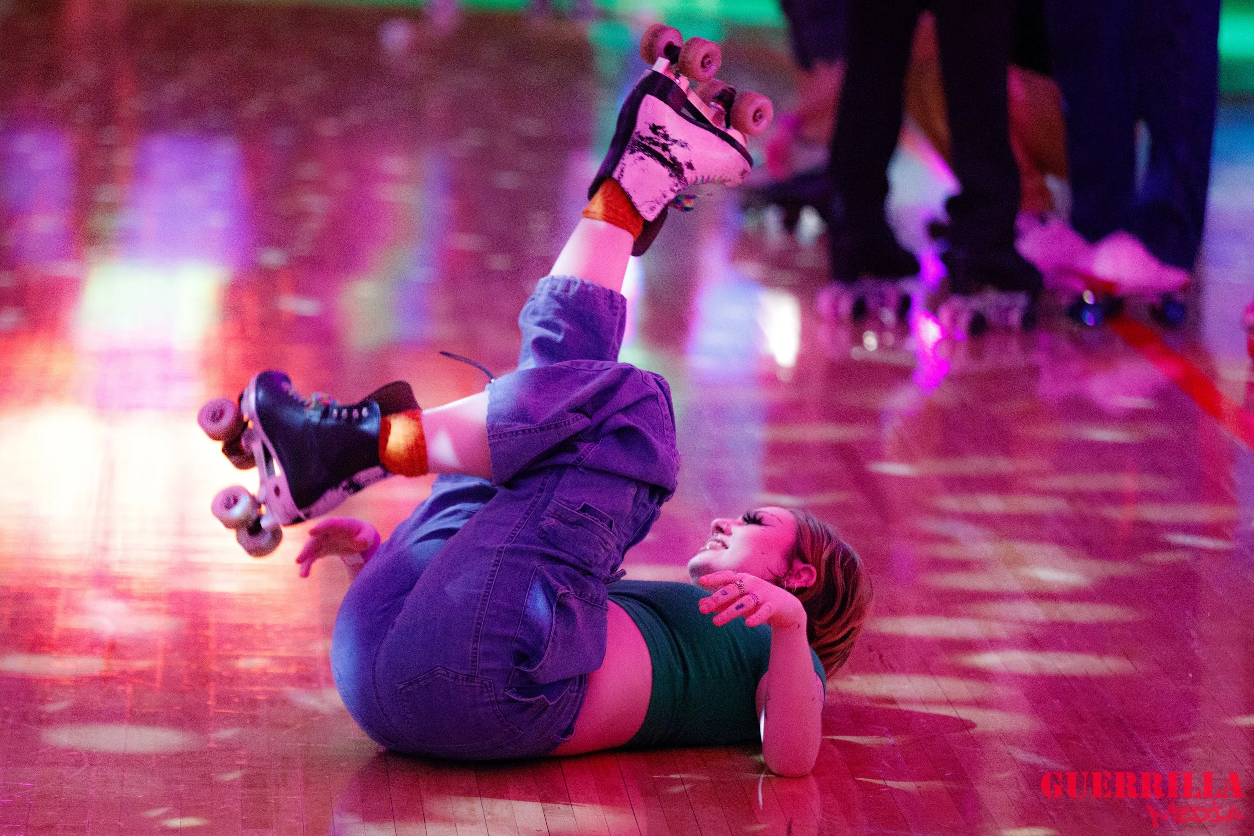 A person with short hair roller skating on a wooden floor, lying on their back with their legs raised and smiling. Neon lights illuminate the scene, and other roller skaters are visible in the background.