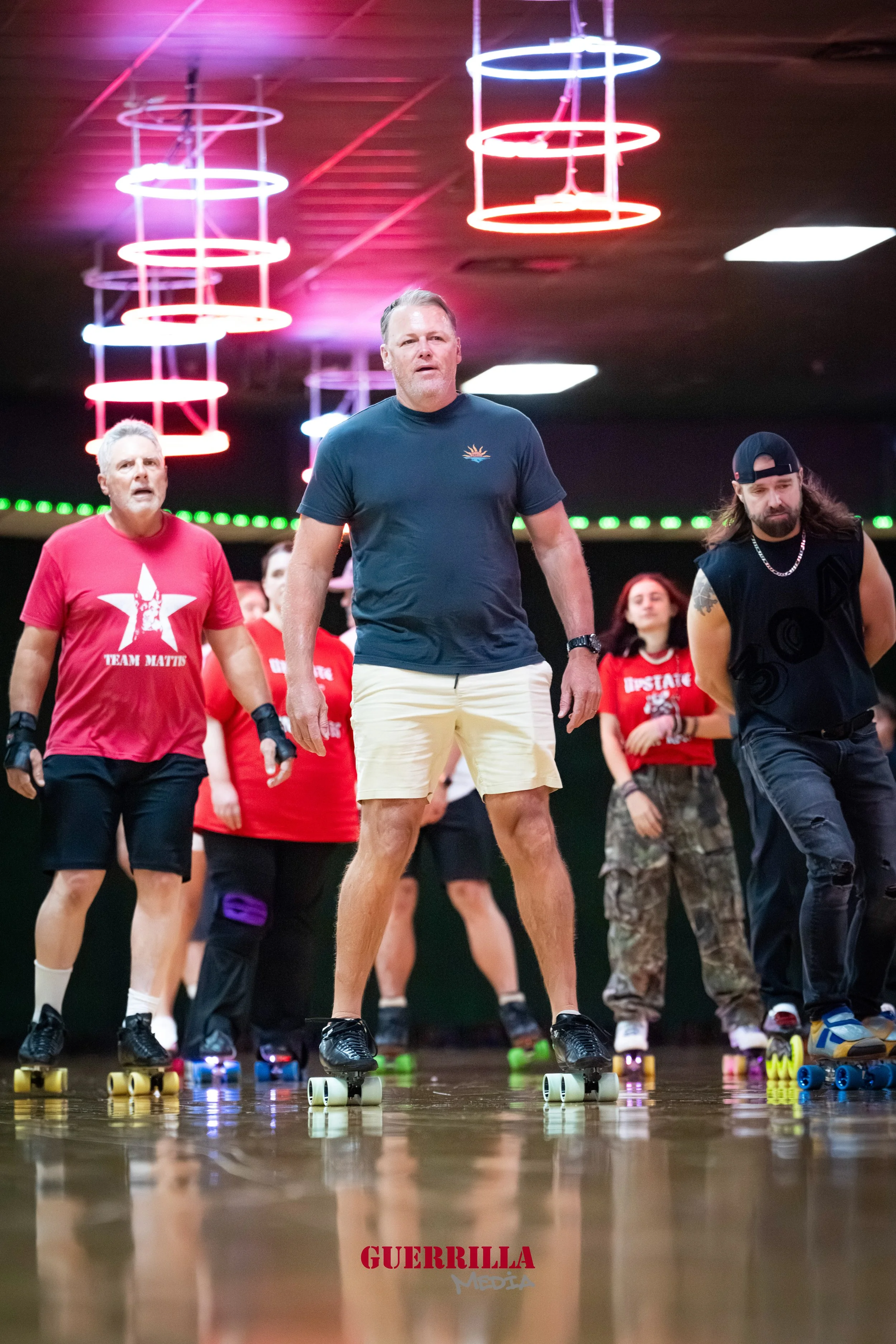 People roller skating indoors, including a man in a blue t-shirt and white shorts, and others in red t-shirts and casual attire, on a wooden rink with neon lighting.