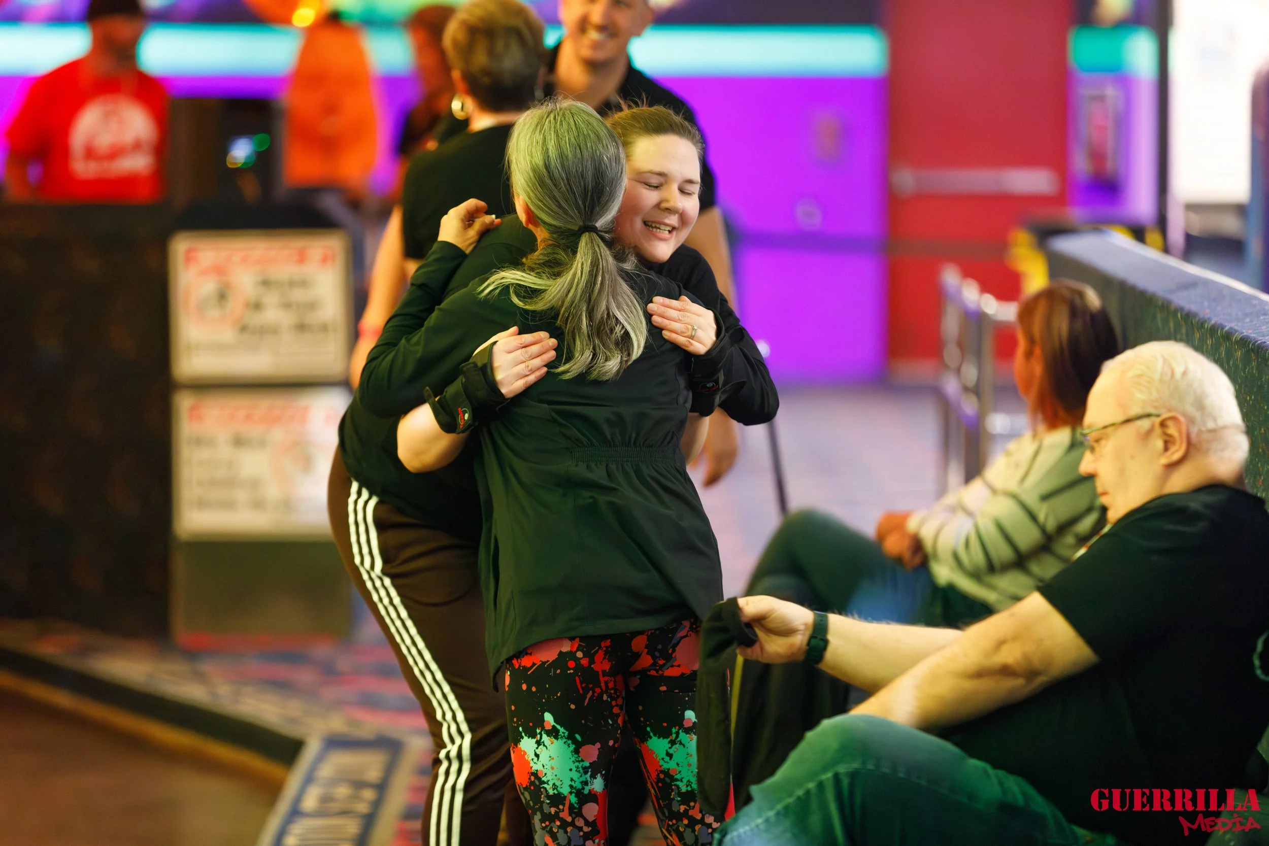 Two women hugging and smiling inside a lively entertainment venue with colorful neon lights, while others sit and stand nearby.