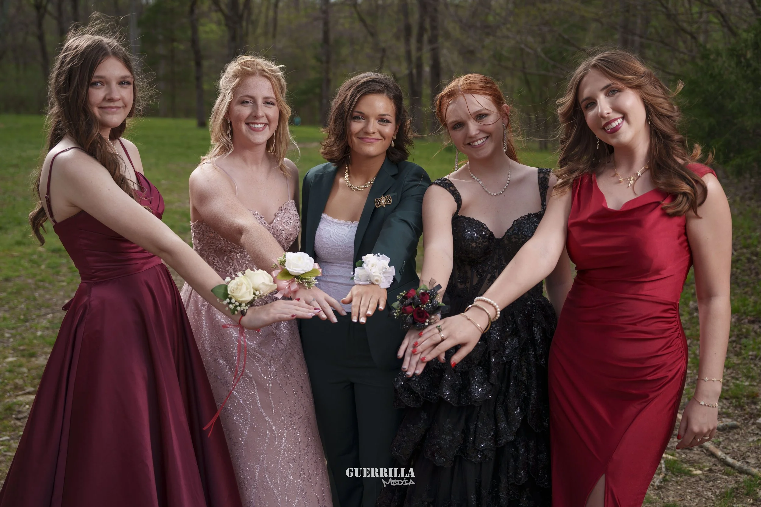Five women in formal dresses standing outdoors in a line with their hands stacked together, smiling, with trees in the background.