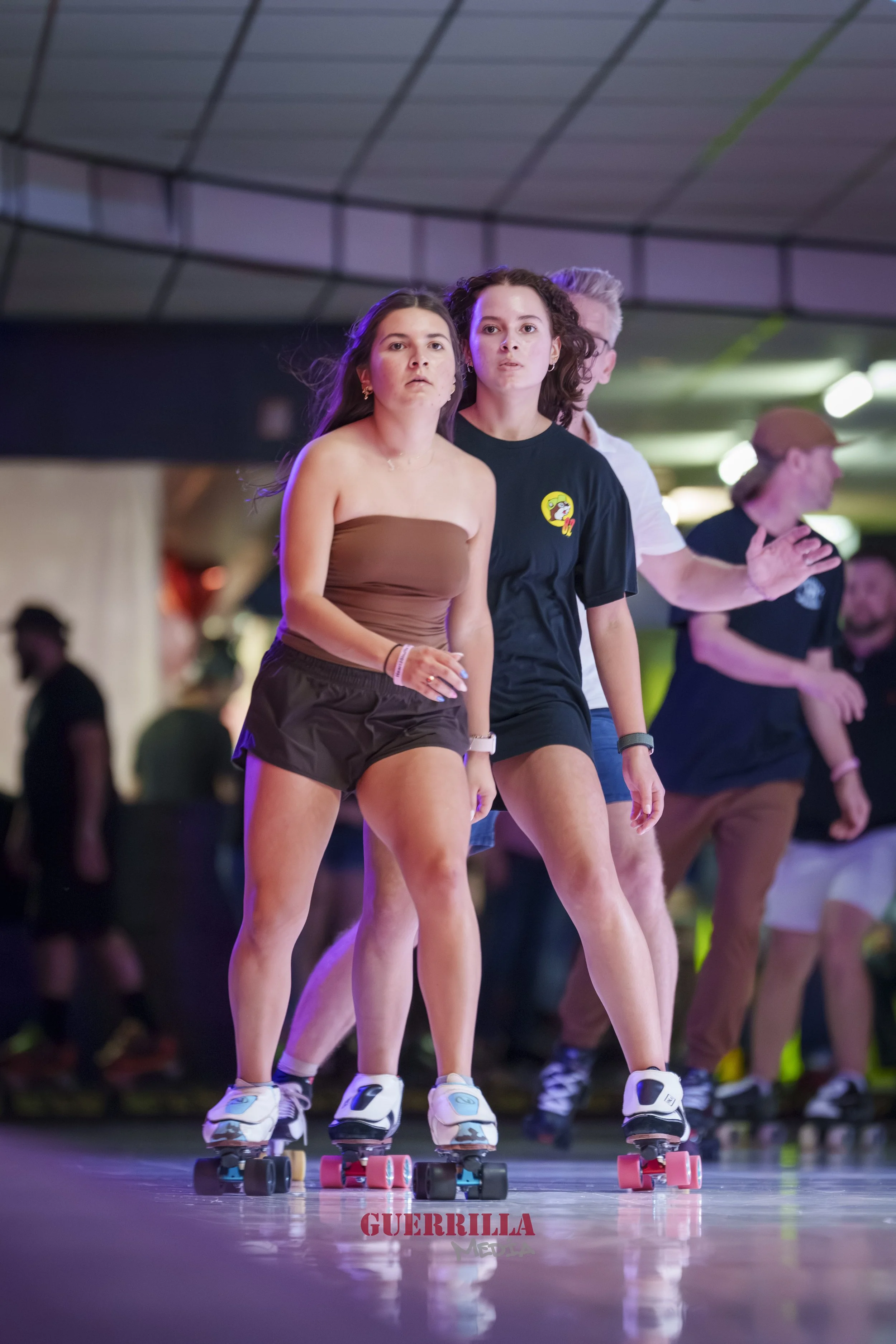 Two young women roller skating indoors, with a group of people in the background, under a ceiling with geometric panels.