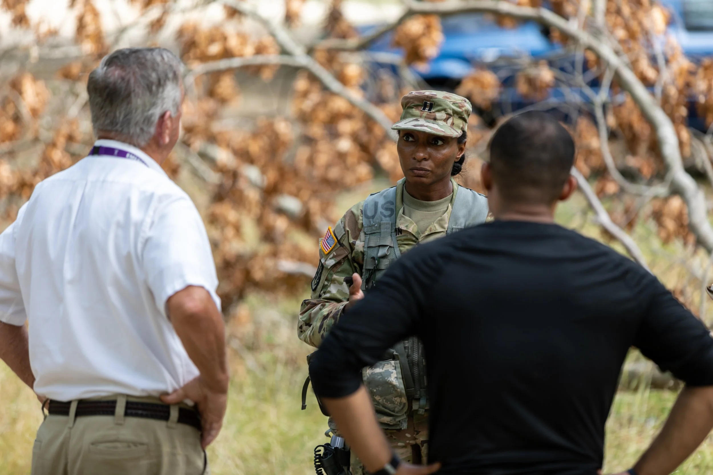 A soldier in camouflage uniform with a US flag patch talking to two men outdoors, with brown autumn leaves on trees in the background.