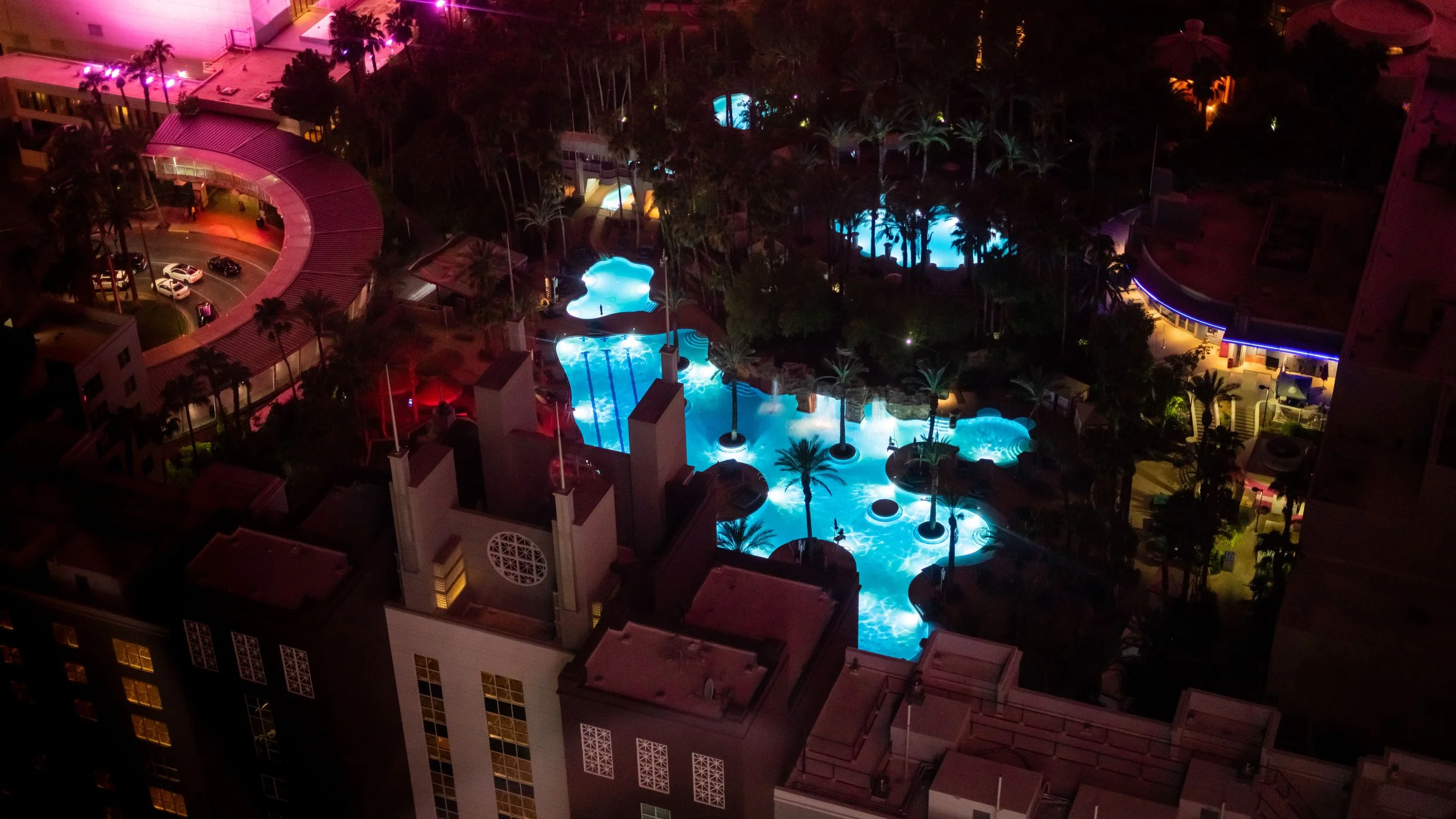 An aerial view of a hotel pool area at night, illuminated in blue and other vibrant colors, surrounded by palm trees and multi-story hotel buildings.