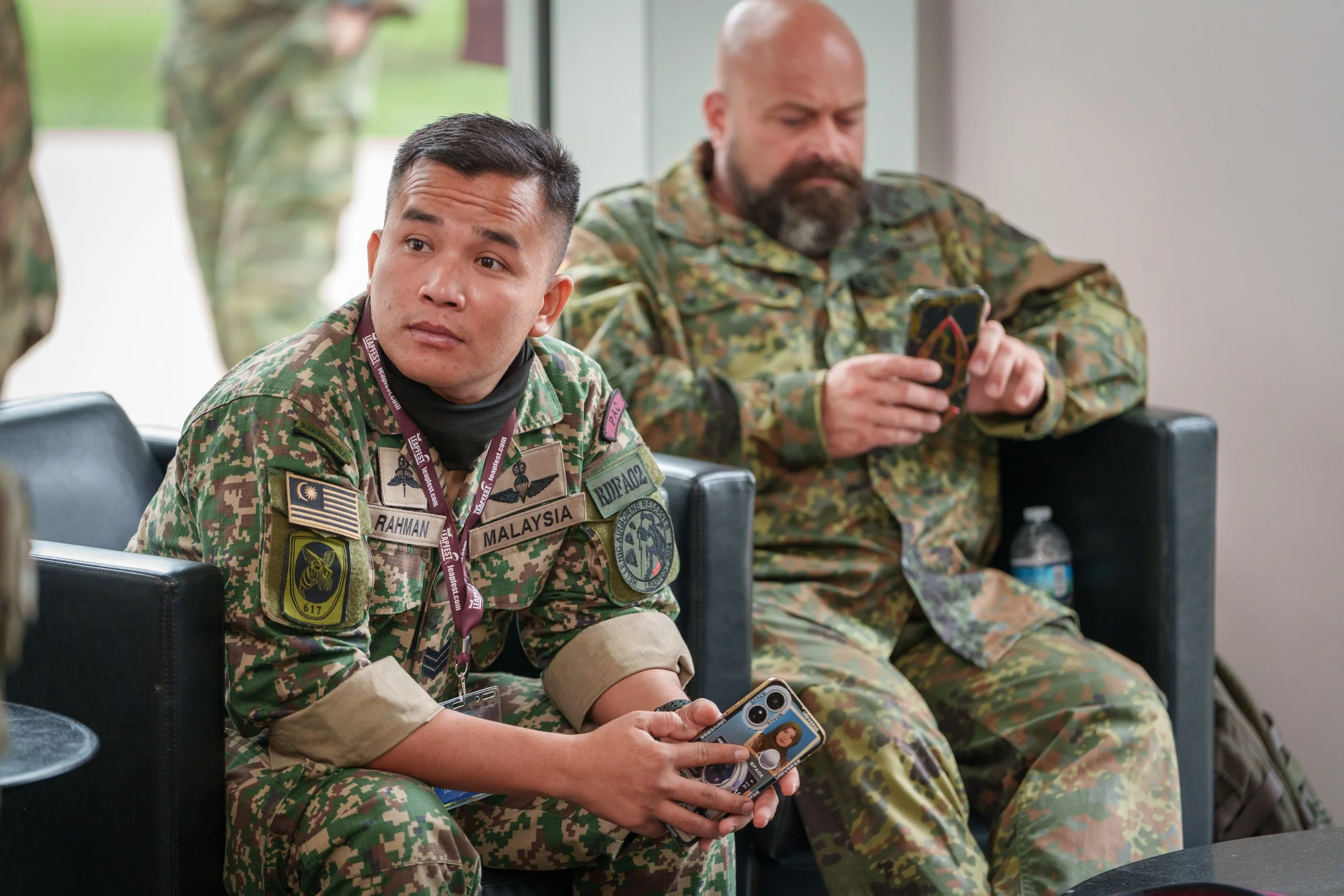 Two military personnel in camouflage uniforms sitting on black chairs, one is a young man with short dark hair holding a smartphone and looking to his left, the other is an older man with a beard who is looking at his smartphone. There is a water bot