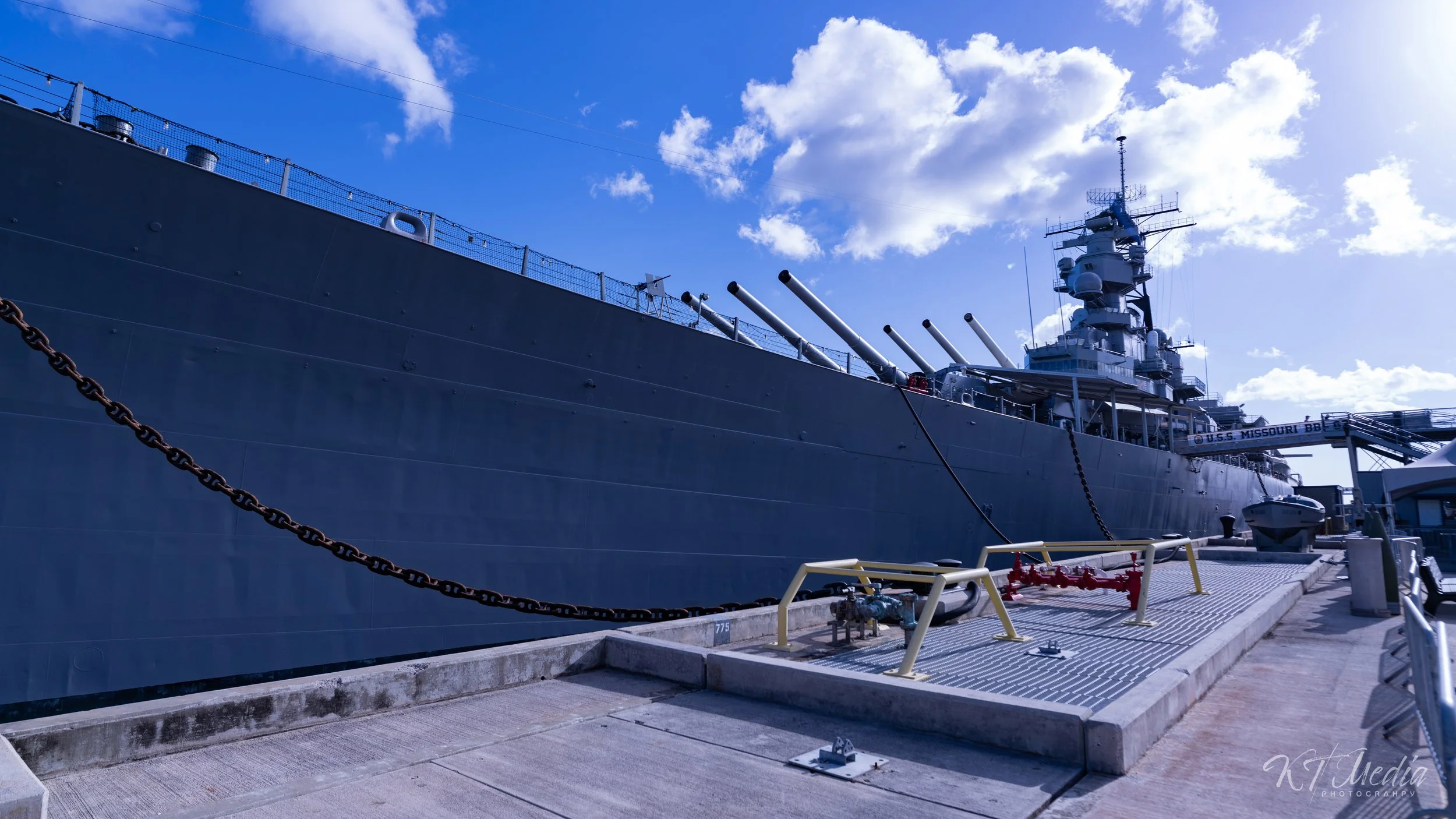 A large military battleship docked at a harbor, with a clear blue sky and scattered clouds above.