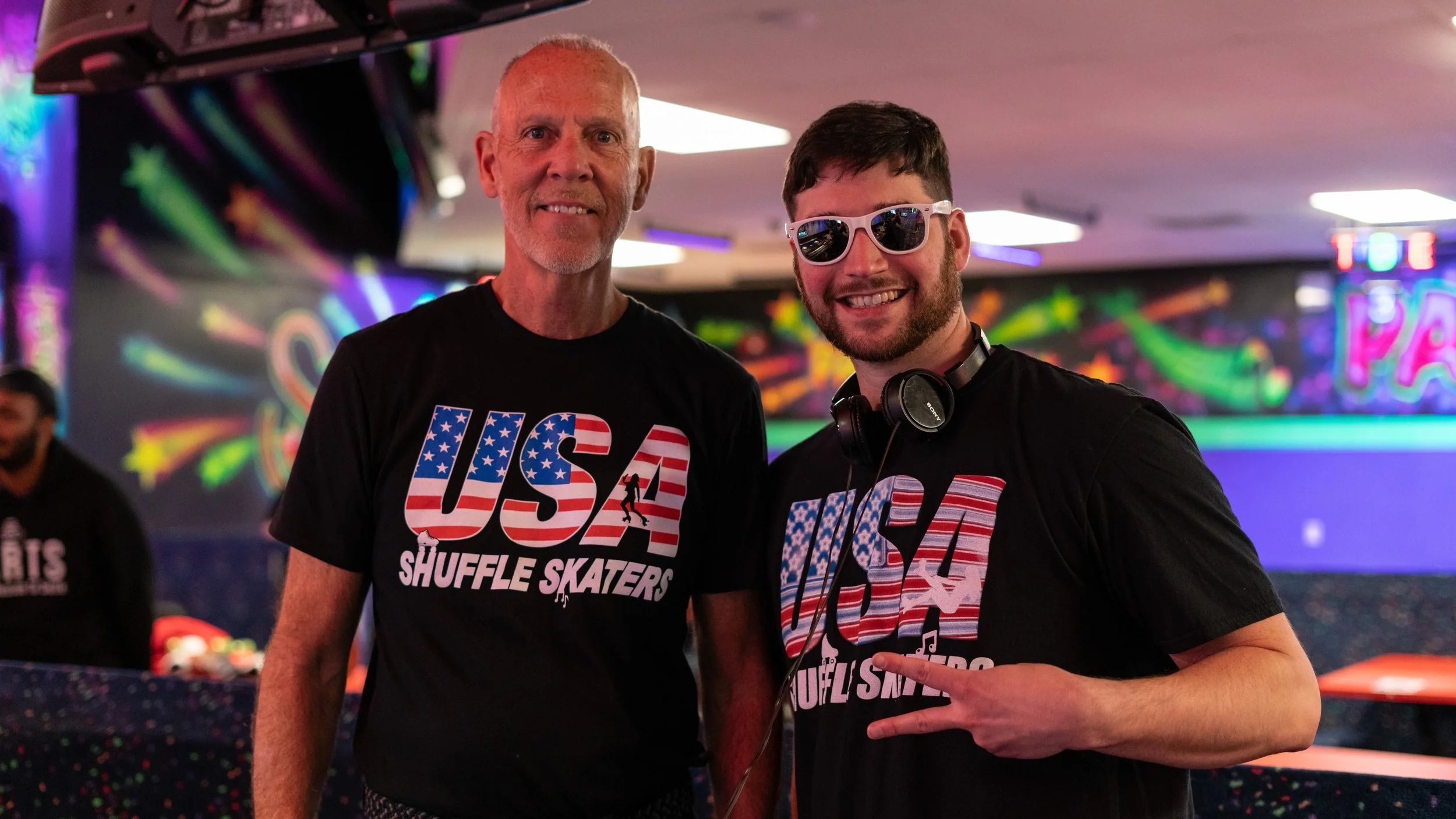 Two men at an indoor roller skating rink, smiling for the camera. Both are wearing black T-shirts with 'USA Shuffle Skaters' in patriotic colors. The background features colorful, neon graffiti-style wall art and other skaters.