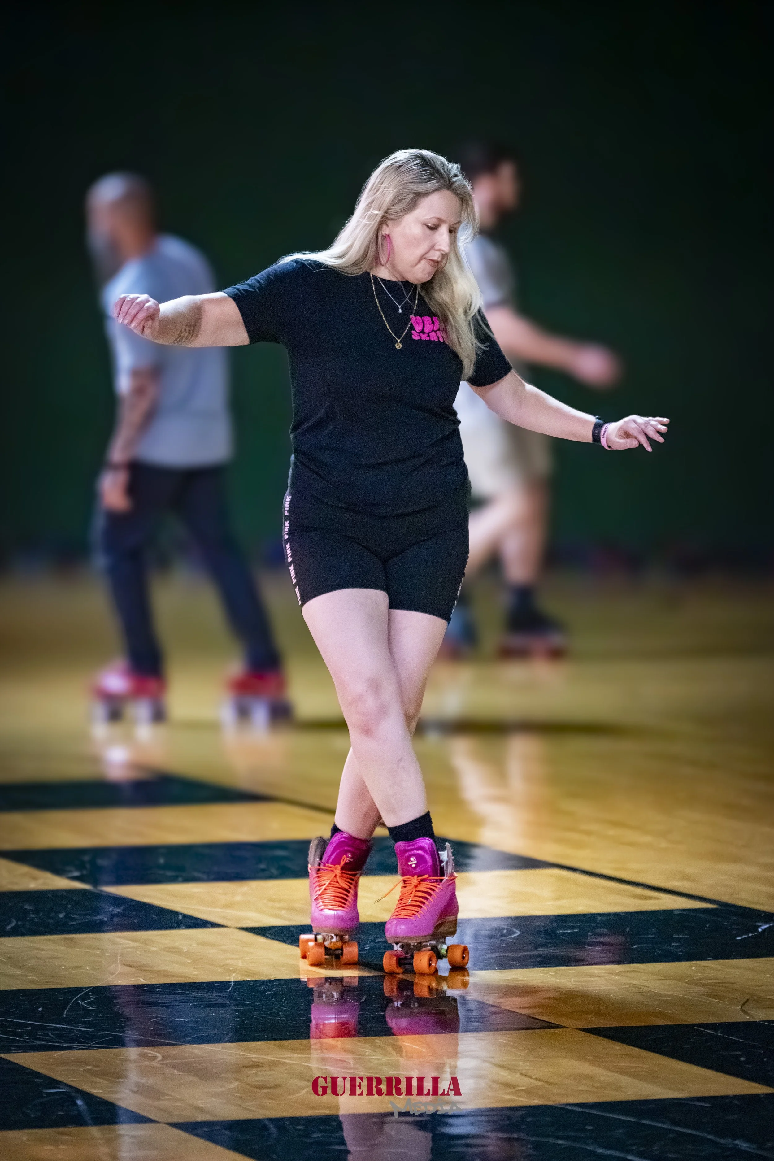 A woman roller skating indoors on a black and yellow checkered floor, with two other skaters in the background.