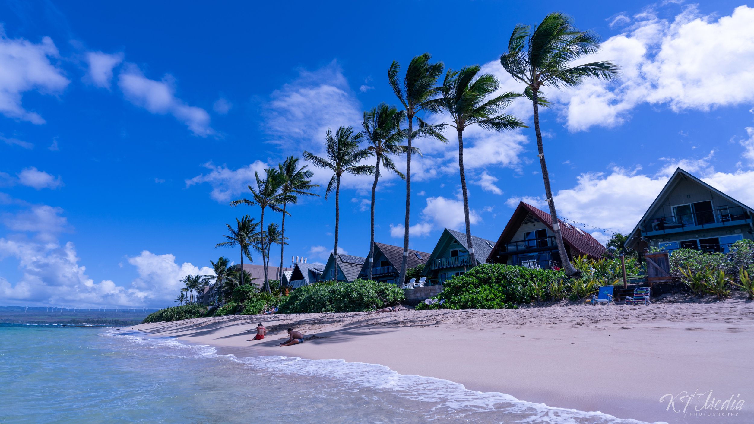 Beach scene with white sand, clear water, palm trees, and several houses with balconies facing the ocean under a blue sky with clouds.