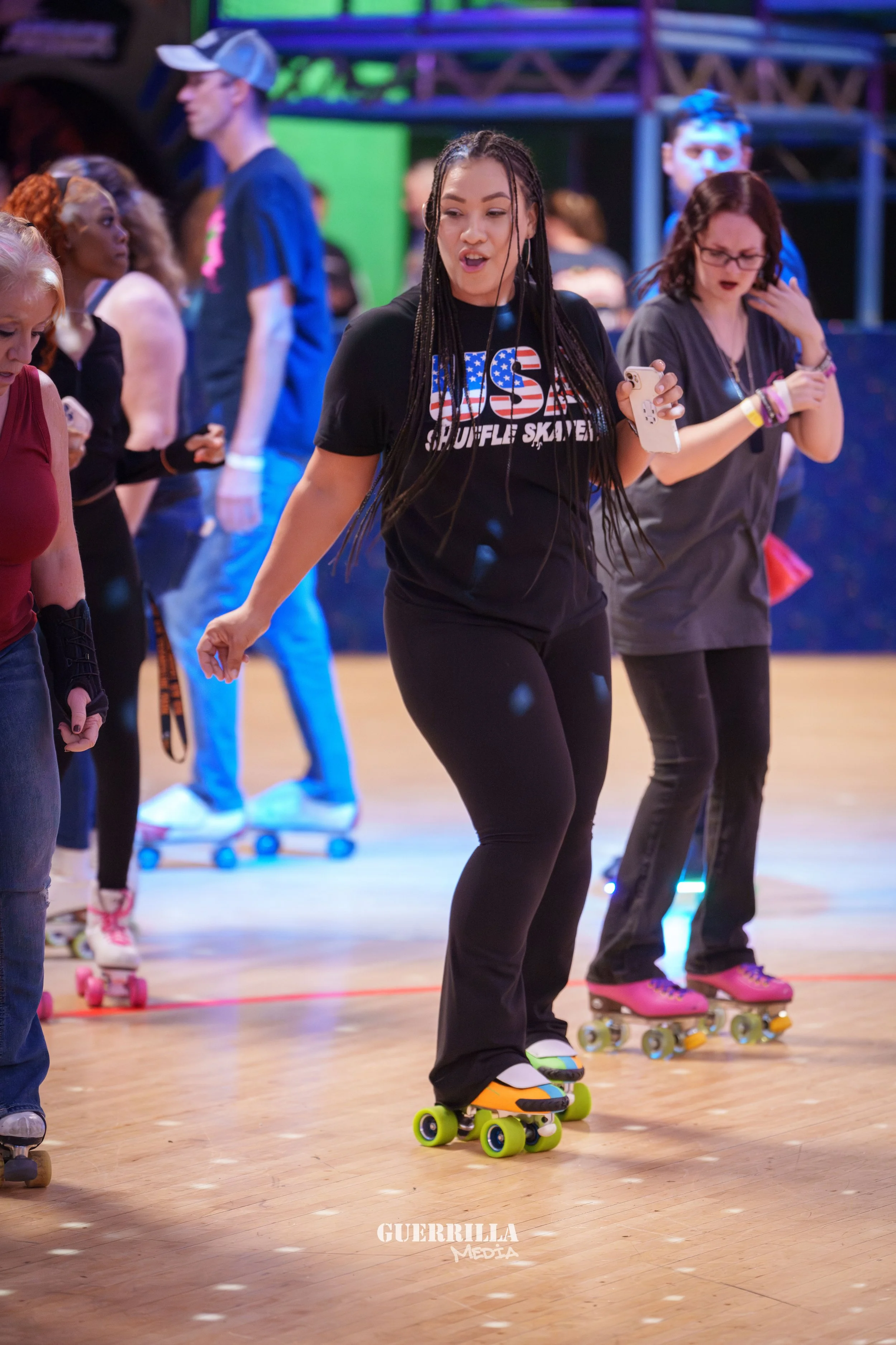 A group of women roller skating on a wooden floor, with one woman in the center wearing a black t-shirt with USA written on it, holding a phone, and wearing pink roller skates with white wheels.