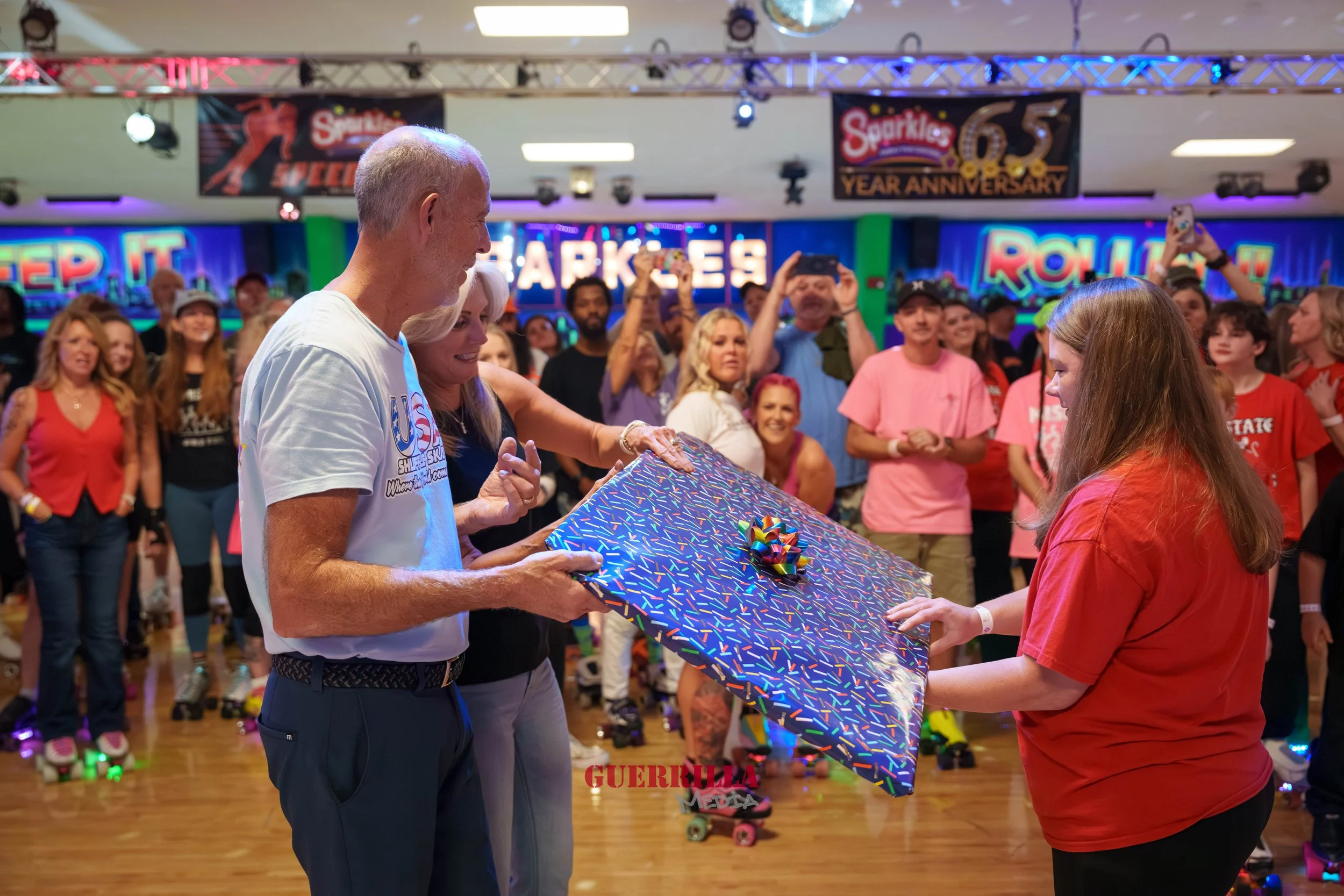 A woman is presenting a large, wrapped gift with a rainbow-colored bow to a woman in a red shirt at a roller skating rink during a celebration, with a crowd watching and taking photos in the background.