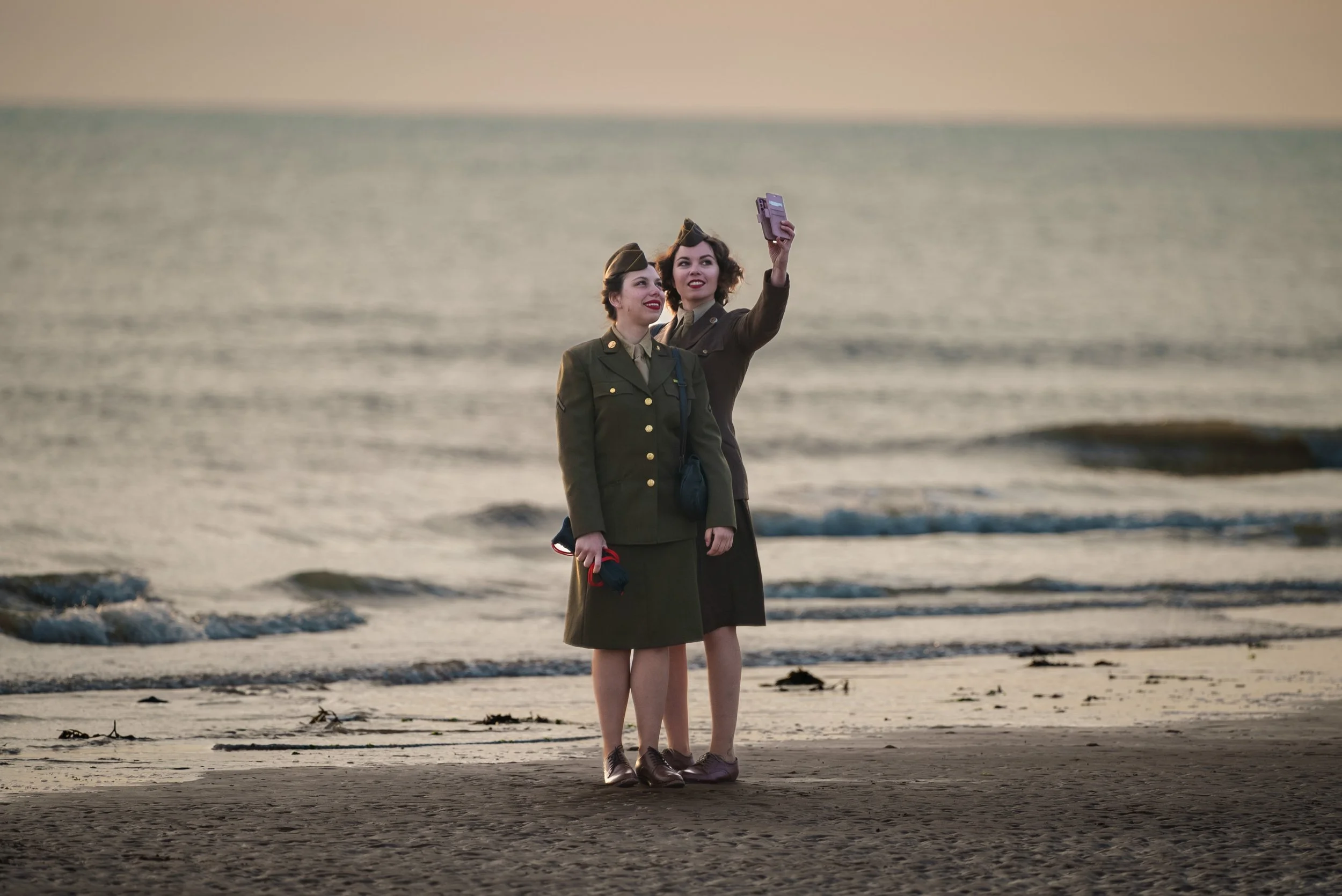 Two women in military uniforms taking a selfie on the beach during sunset.