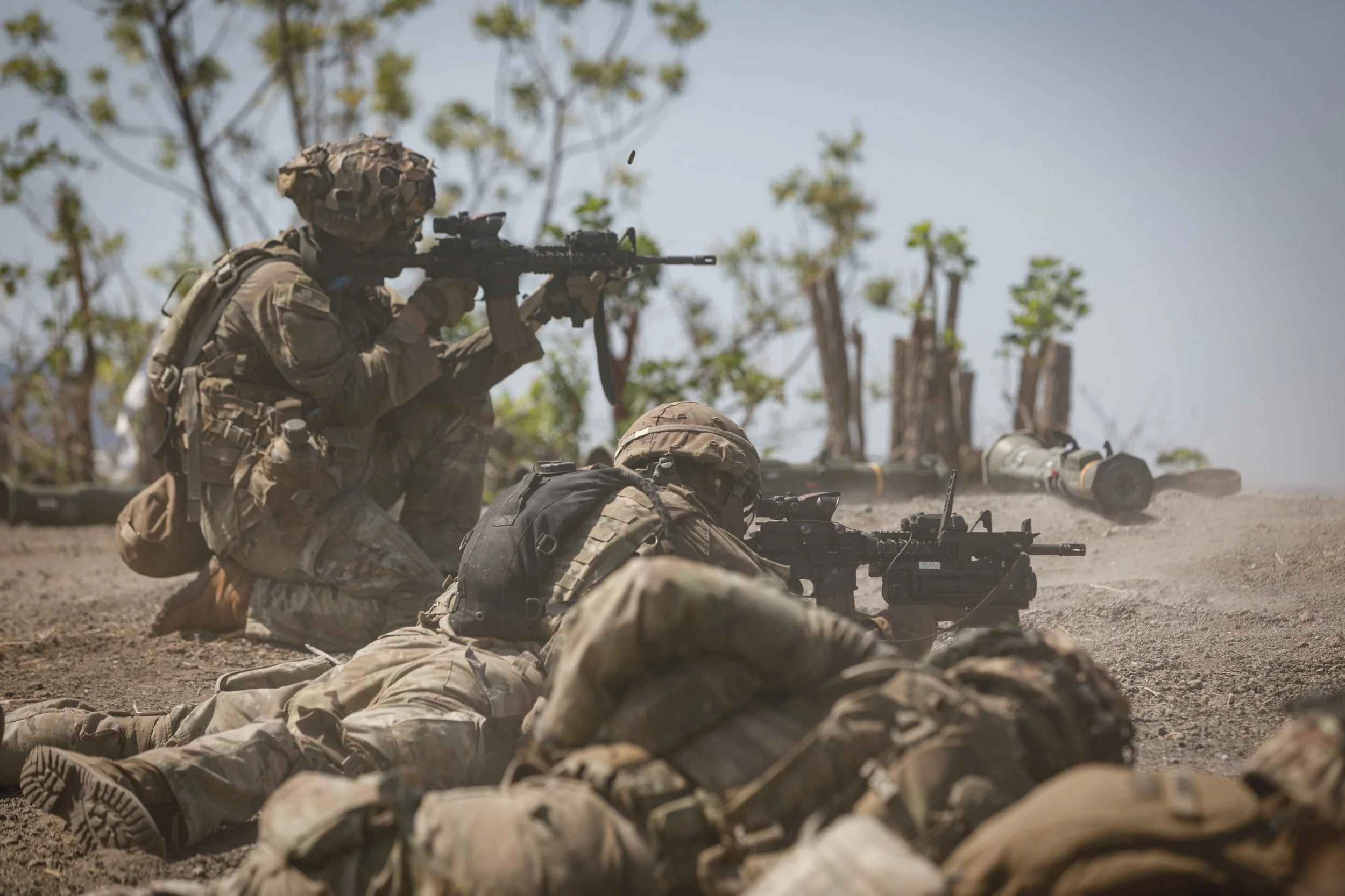 Soldiers in camouflage gear aiming rifles while crouching and lying on the ground during a military operation in a dusty outdoor environment.