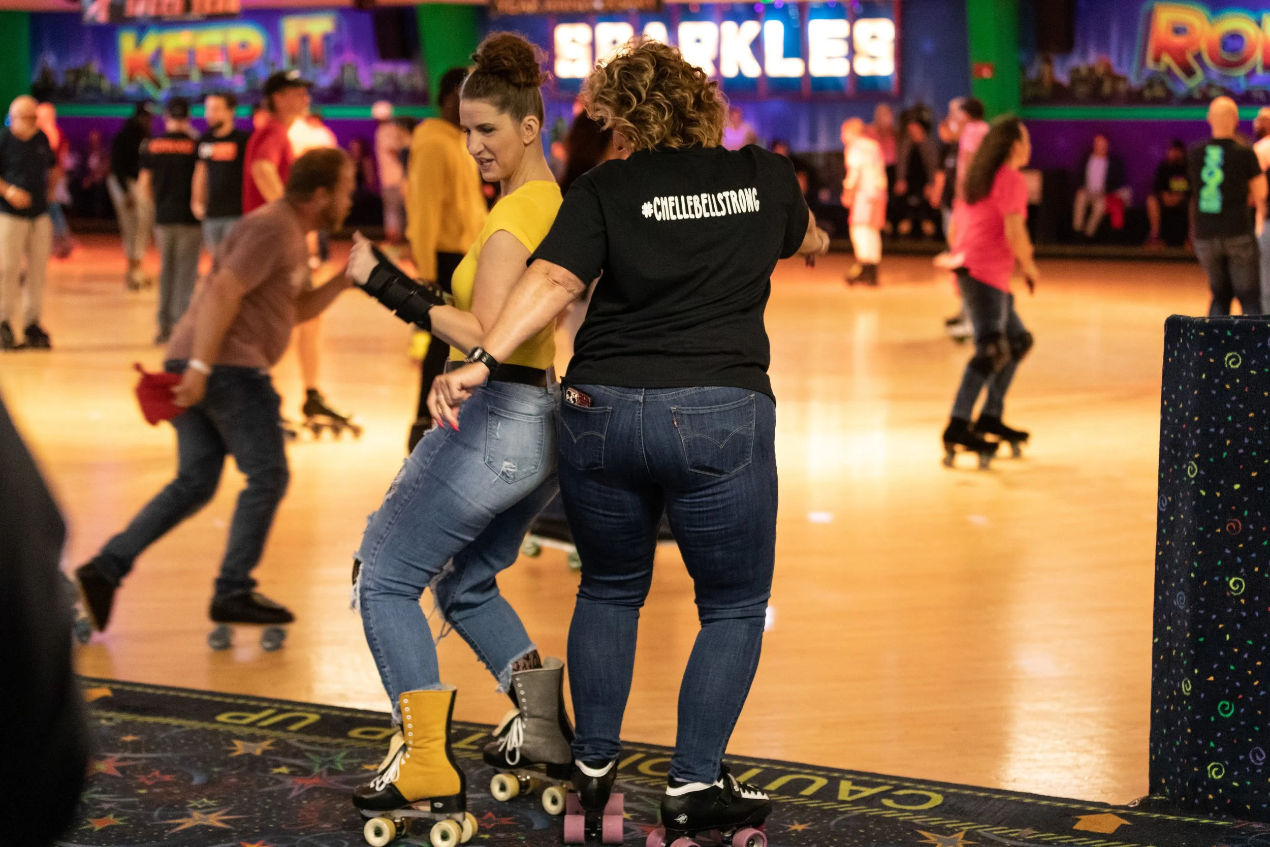 Two women rollerskating at a roller rink, one wearing a black t-shirt with '#CHELLEBELISTRONG' on the back, and the other in a yellow t-shirt.