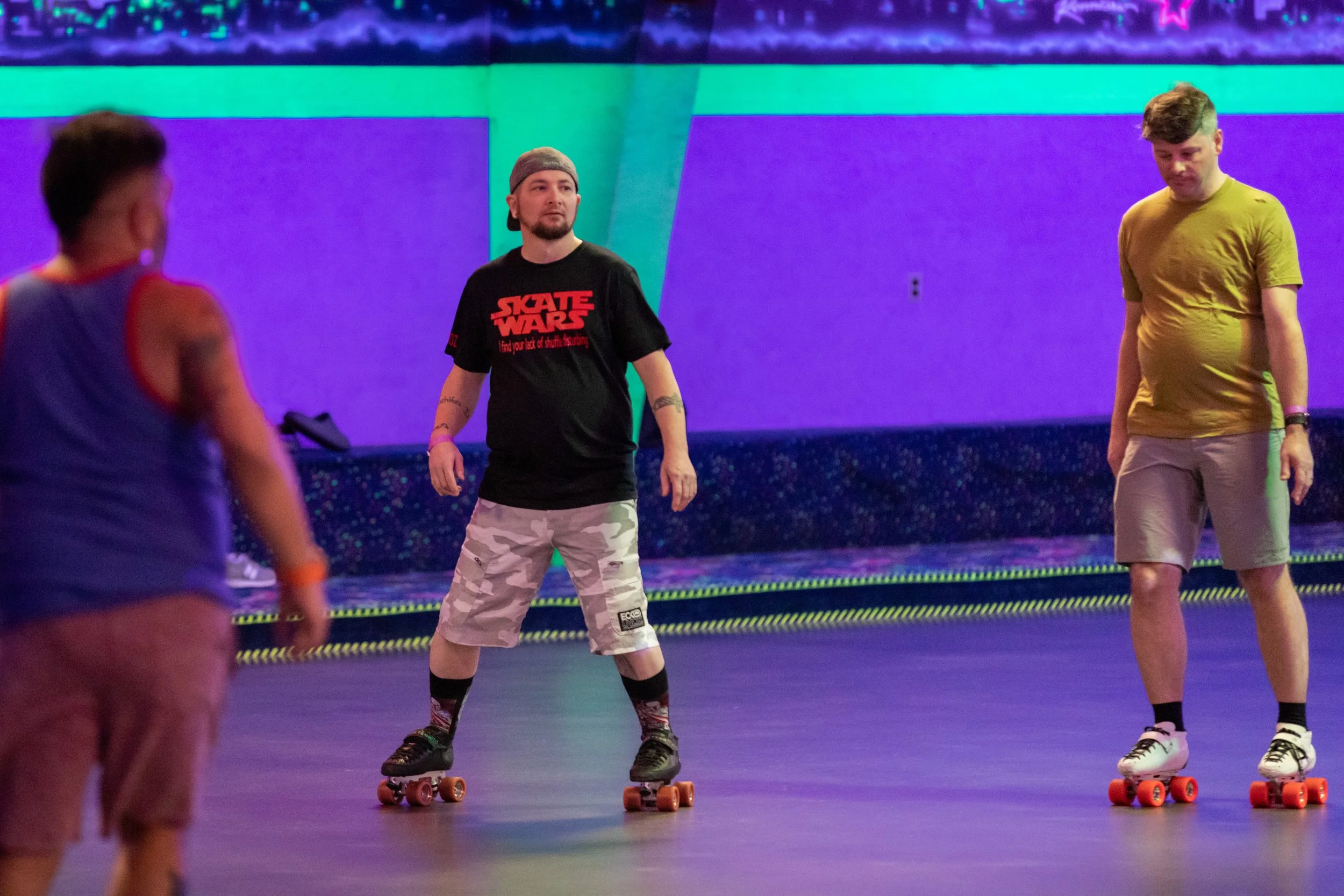 Three men roller skating in an indoor roller rink with colorful lighting. One man in the foreground is slightly blurred, wearing a blue sleeveless shirt. The man in the middle is wearing a black T-shirt with red text, camouflage shorts, and a beanie.