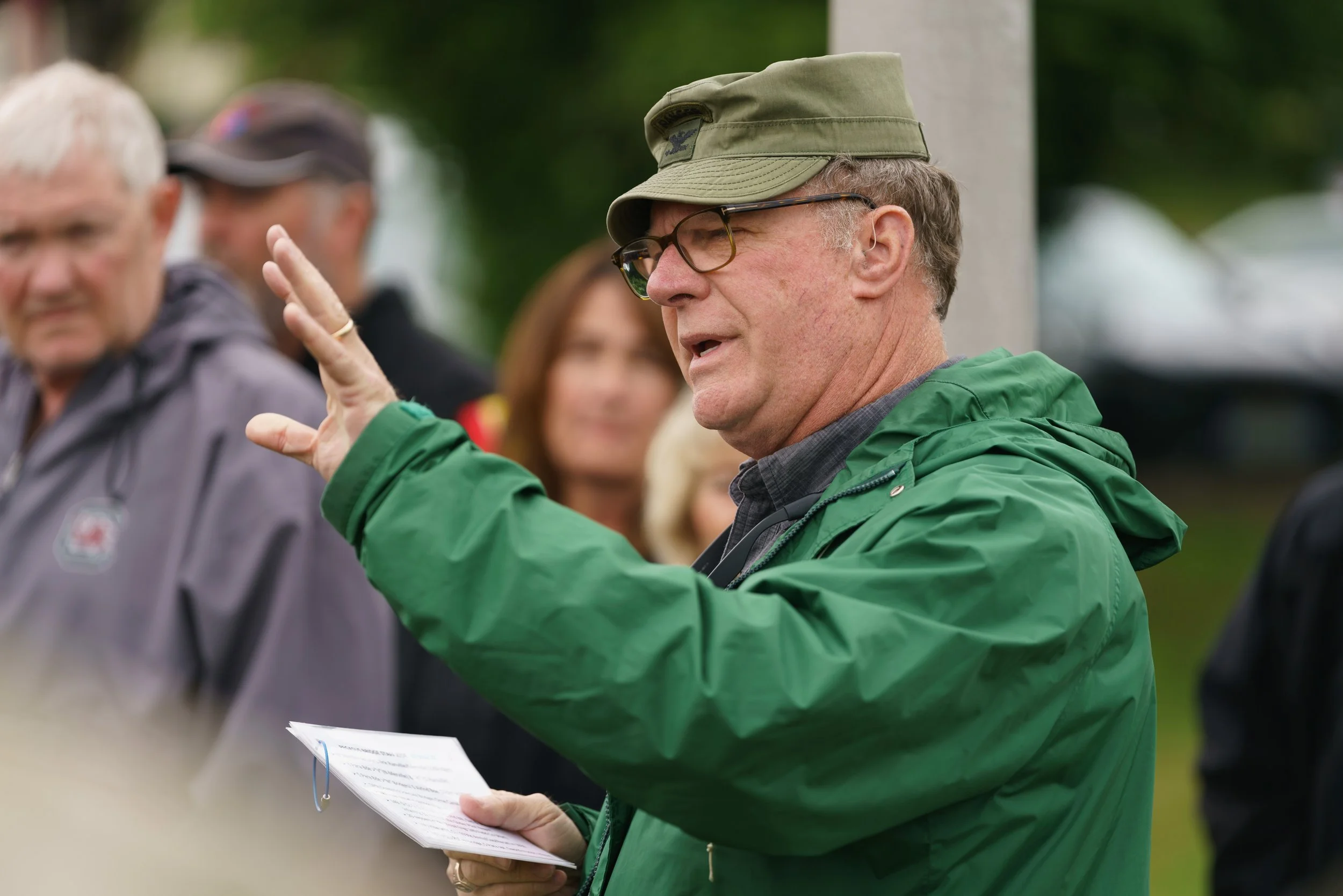 A man wearing a green rain jacket, hat, and glasses, speaking outdoors. He is holding a booklet and gesturing with his hand, with several people standing behind him.