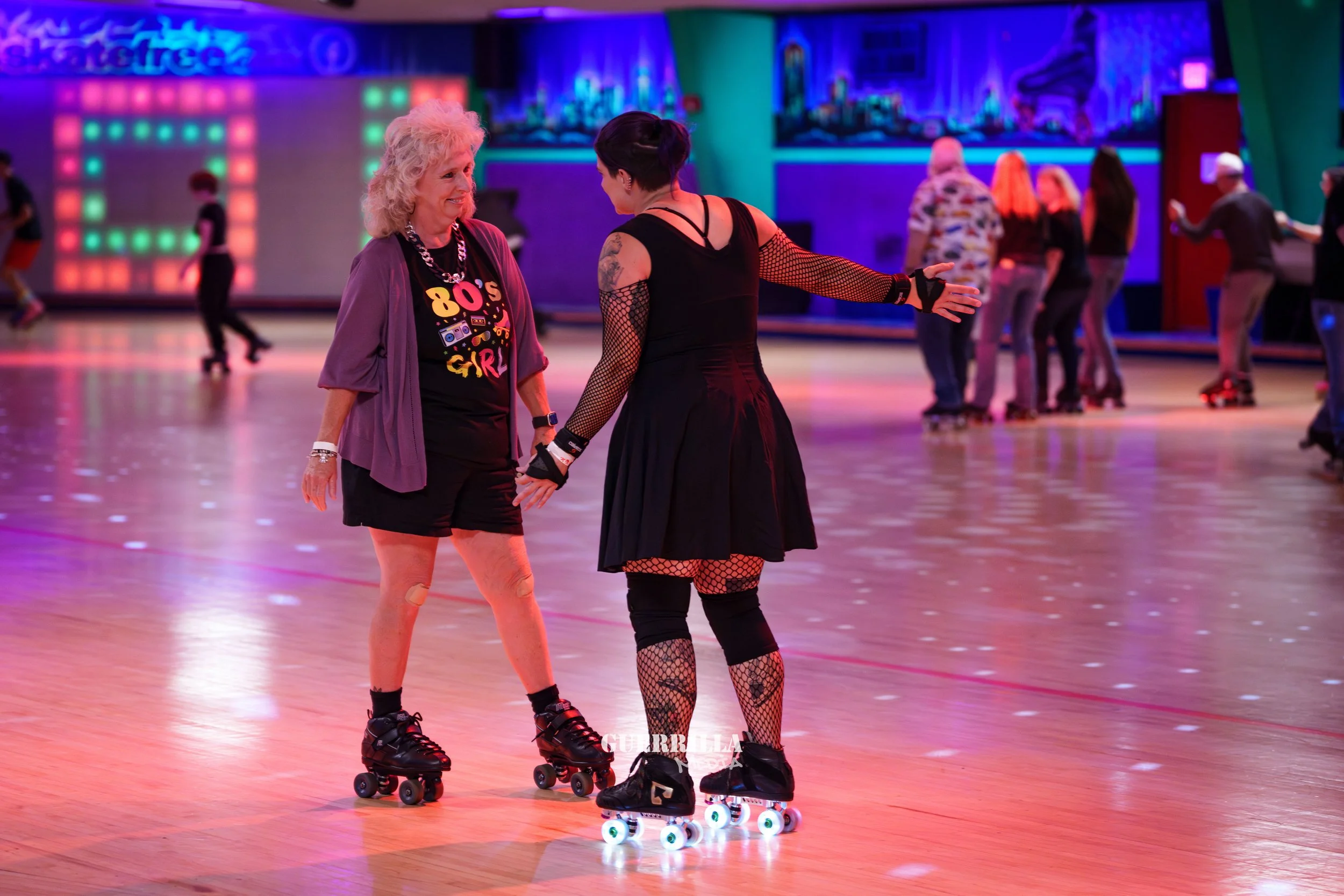 Two women roller skating and holding hands on a roller rink with colorful neon lights and other skaters in the background.