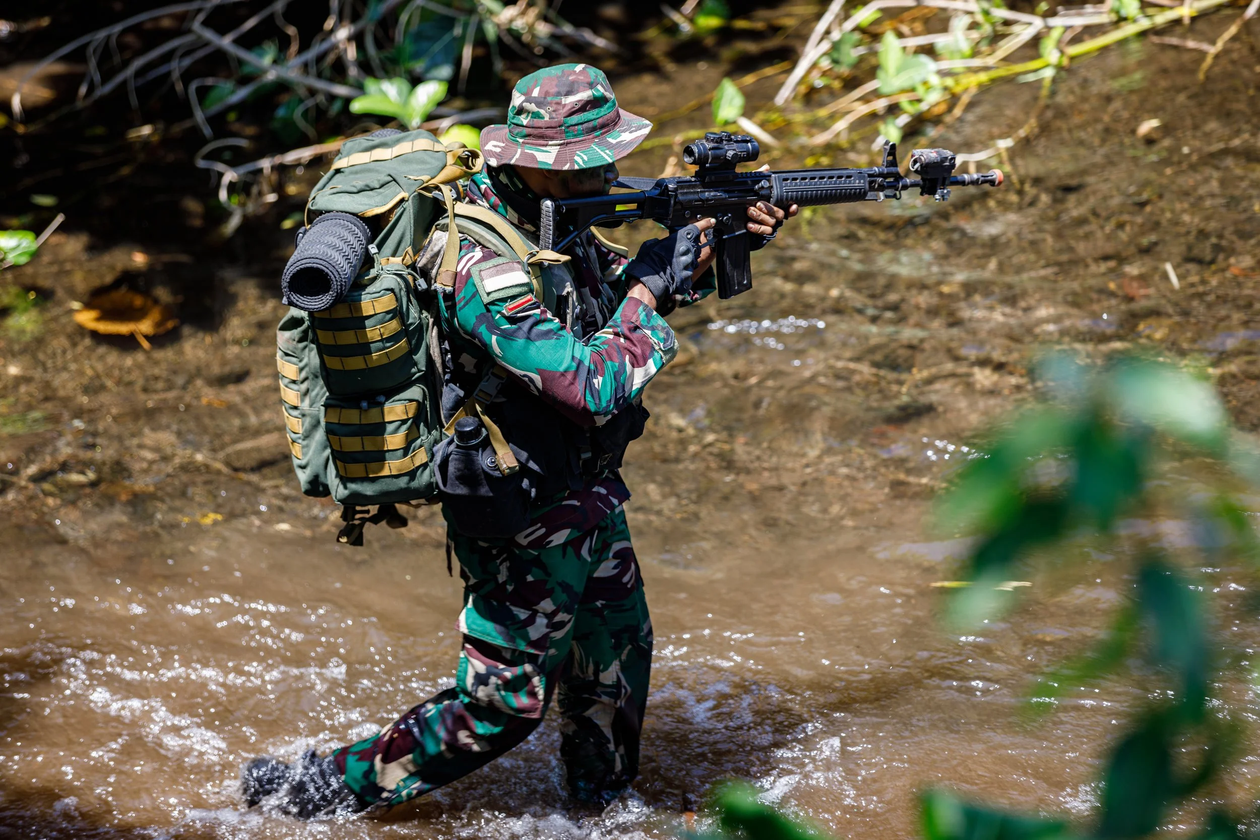 Soldier in camouflage uniform crossing a muddy stream with a rifle and backpack in a forest