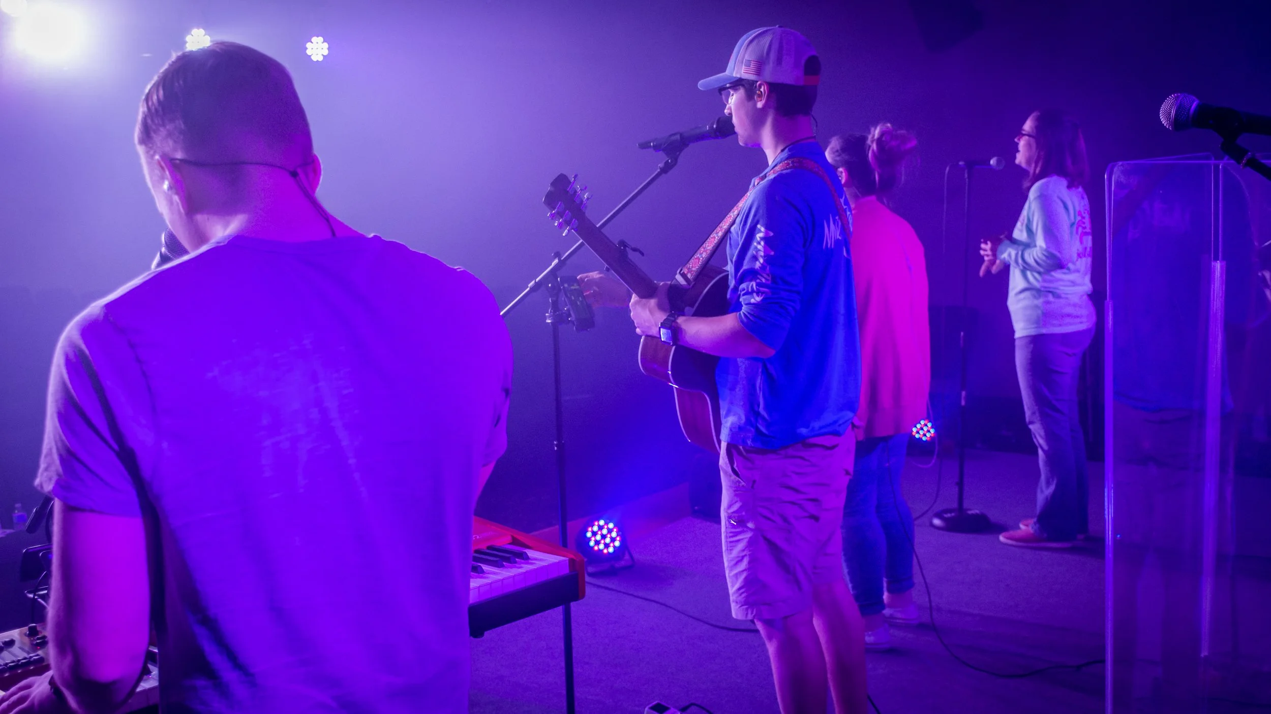 A group of people performing on stage with purple lighting, including a man playing keyboard, a young man singing into a microphone while playing guitar, and several women singing or speaking into microphones.