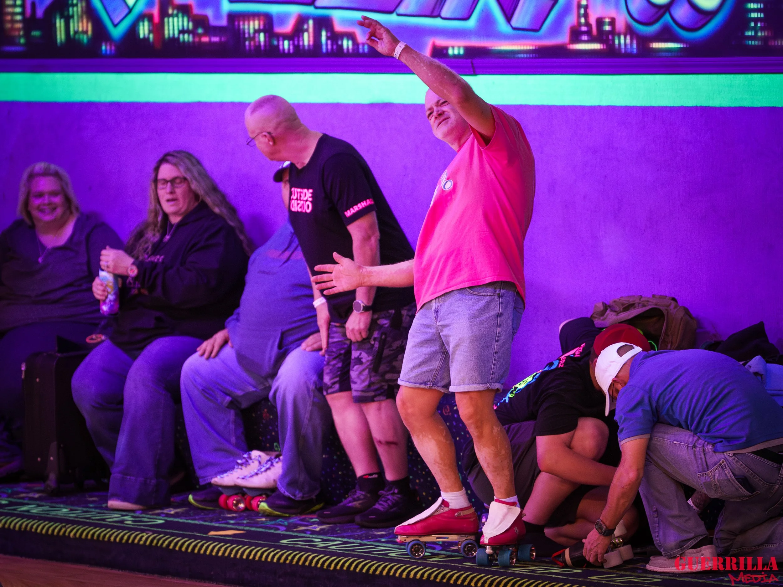 People sitting and standing on a bench under colorful neon lighting, some with roller skates and gear, others holding drinks, in a lively indoor setting.