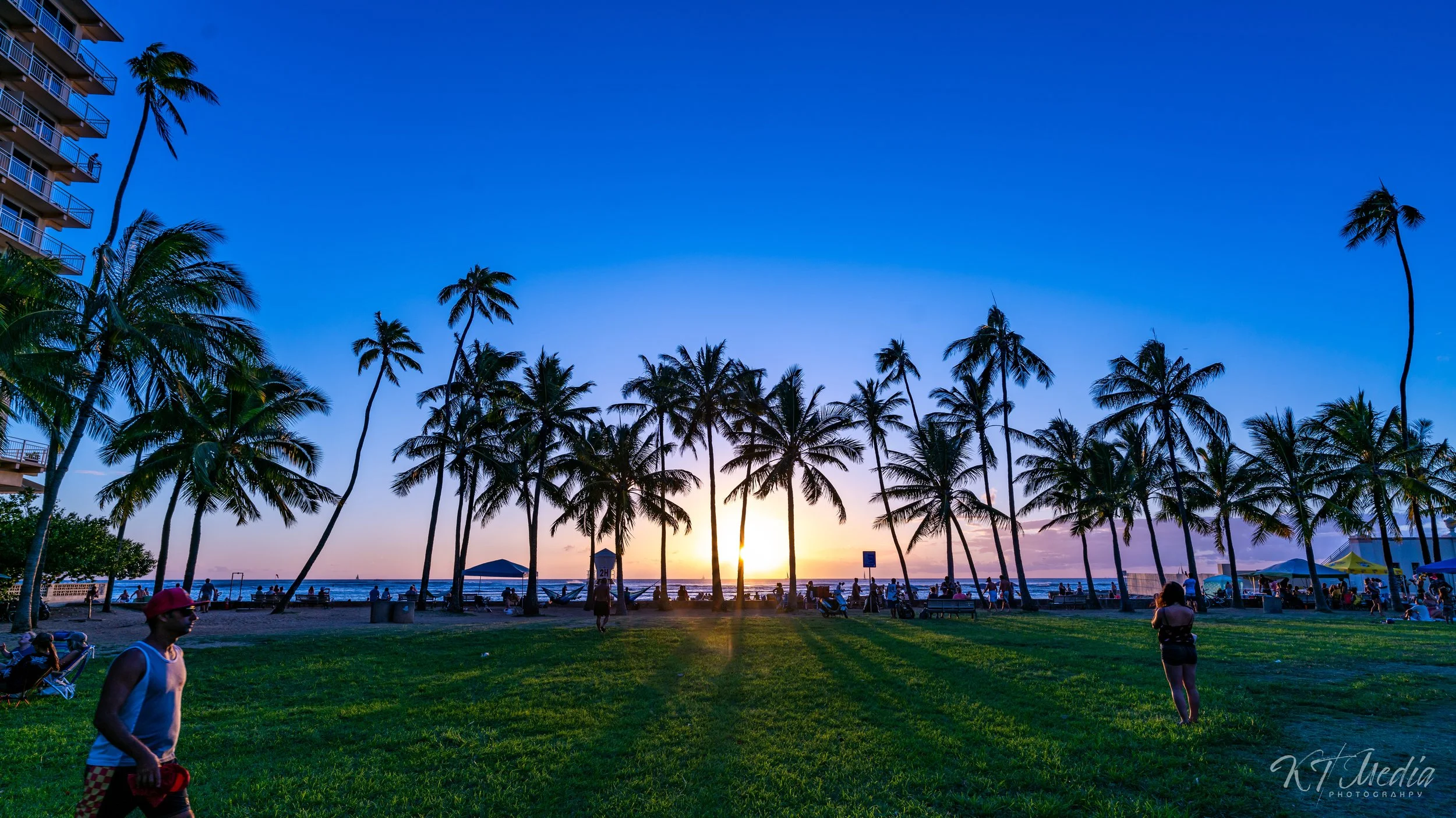 A sunset over a beach park with palm trees, people, and a building visible on the left, in Miami, Florida.