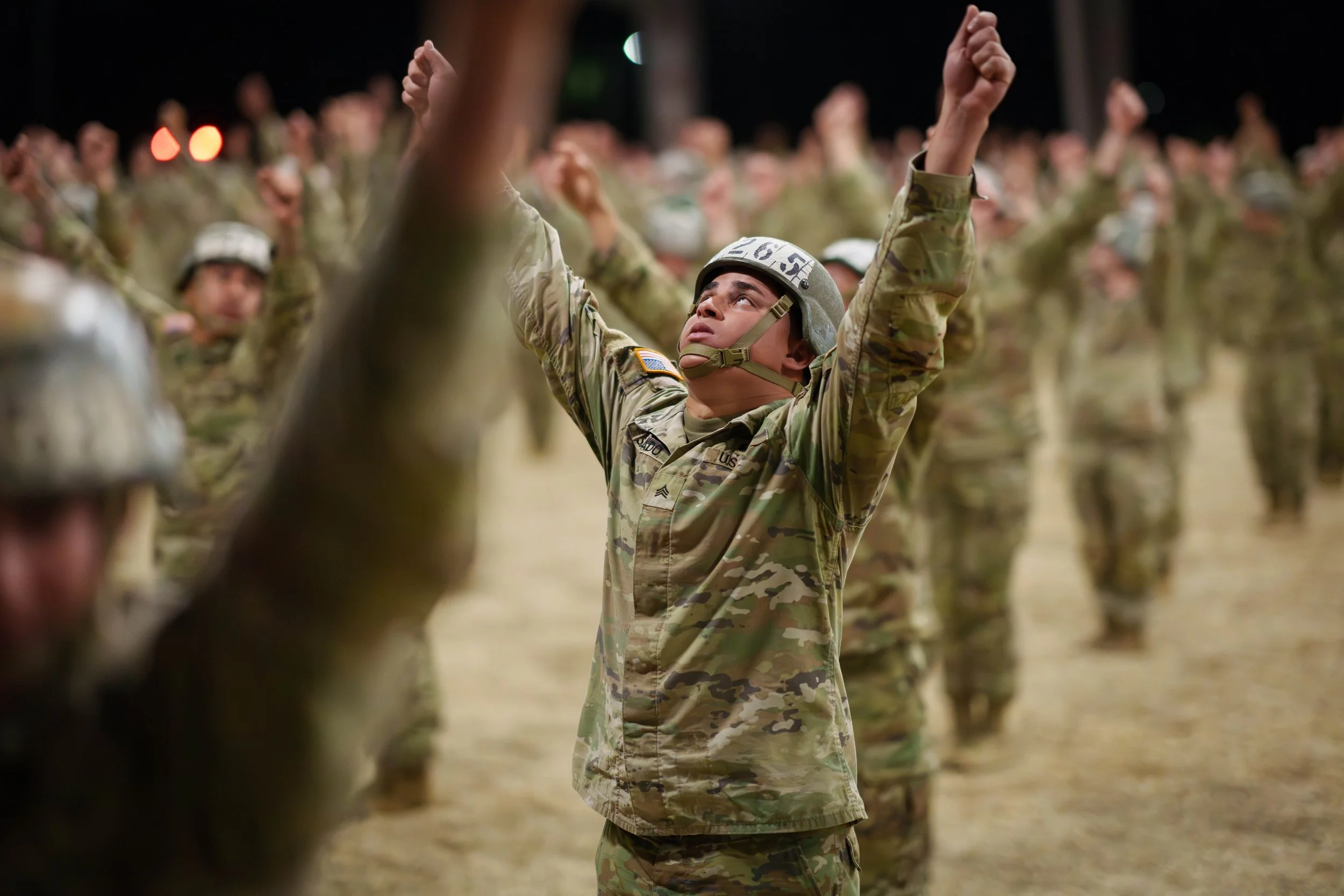 A soldier in camouflage uniform with arms raised, surrounded by other soldiers in similar attire, participating in a military drill or ceremony at night.