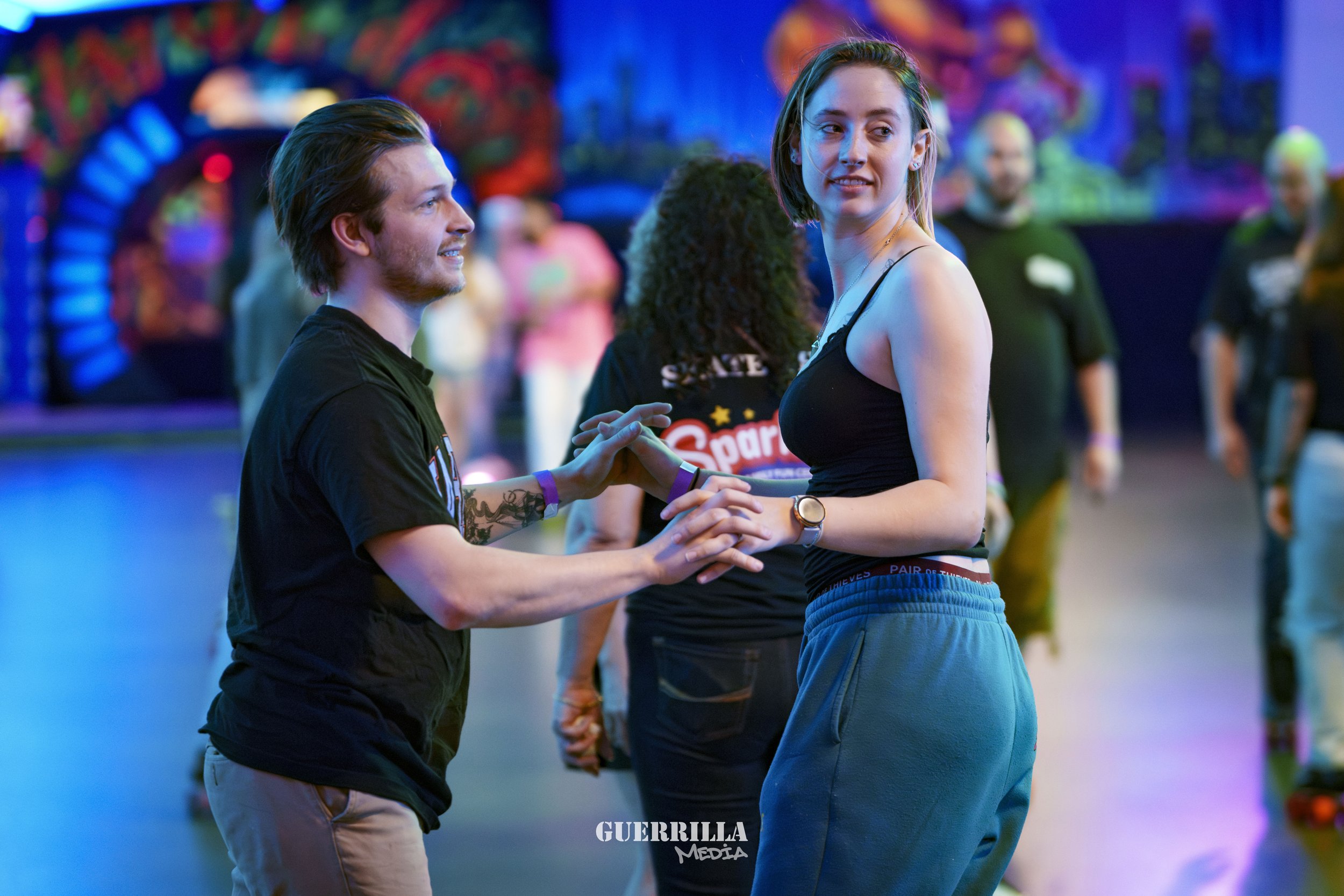 A young man and woman are dancing together at an indoor event with colorful lighting. The man has light skin, brown hair, and is wearing a black t-shirt. The woman has light skin, light brown hair, and is wearing a black tank top and blue sweatpants.