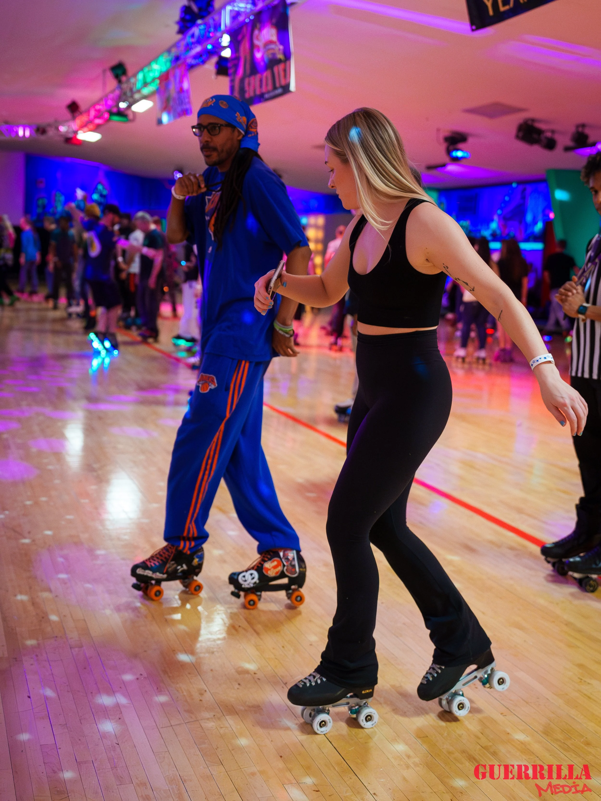 Two women roller skating in a crowded, colorful indoor roller rink with vibrant lights and many people in the background.