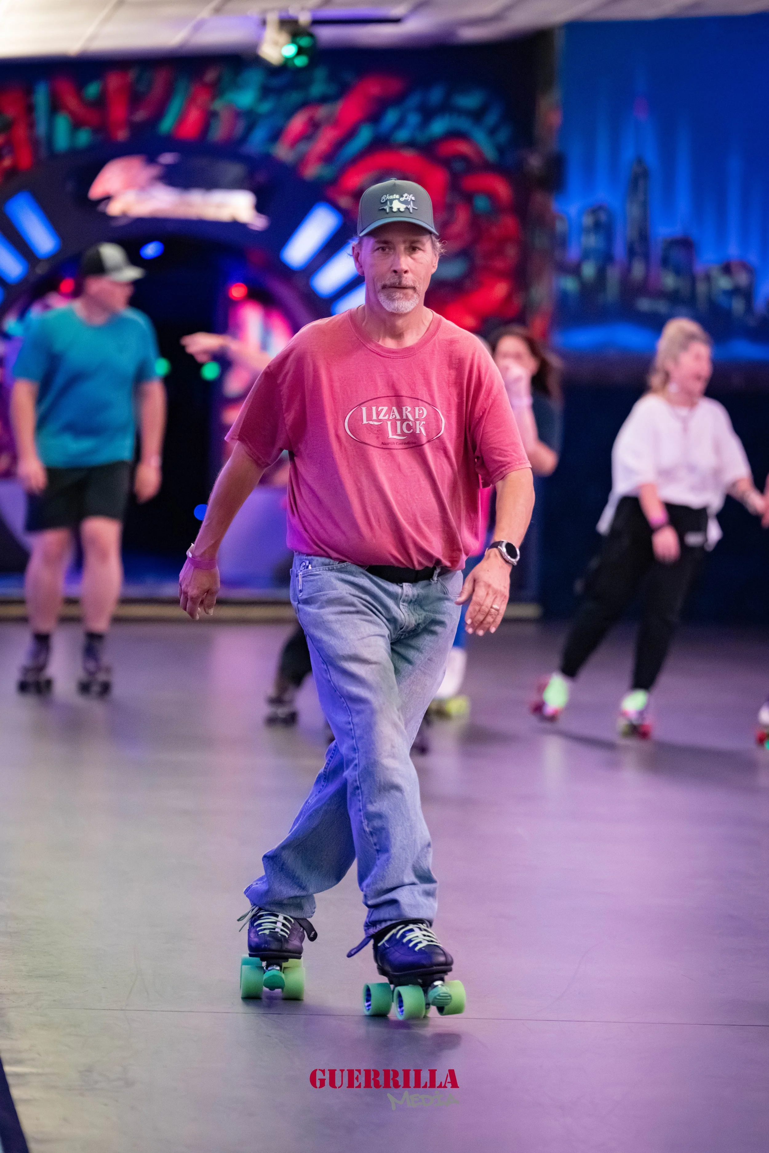 A man roller skating in an indoor roller rink with colorful graffiti art on the wall in the background, wearing a pink shirt, gray jeans, and a gray cap.