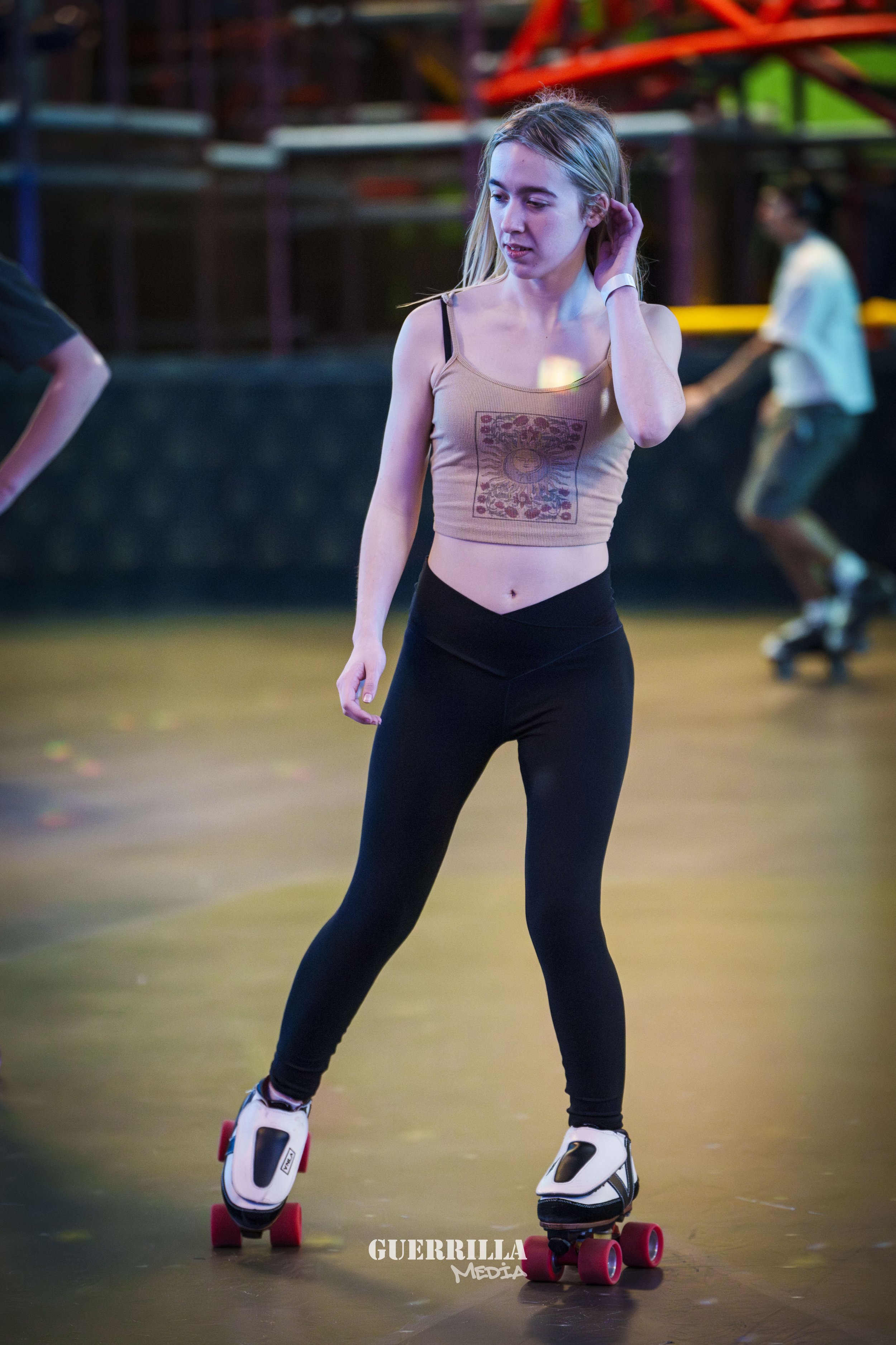 A young woman skating at an indoor roller rink, wearing black leggings and a beige crop top, with other skaters visible in the background.