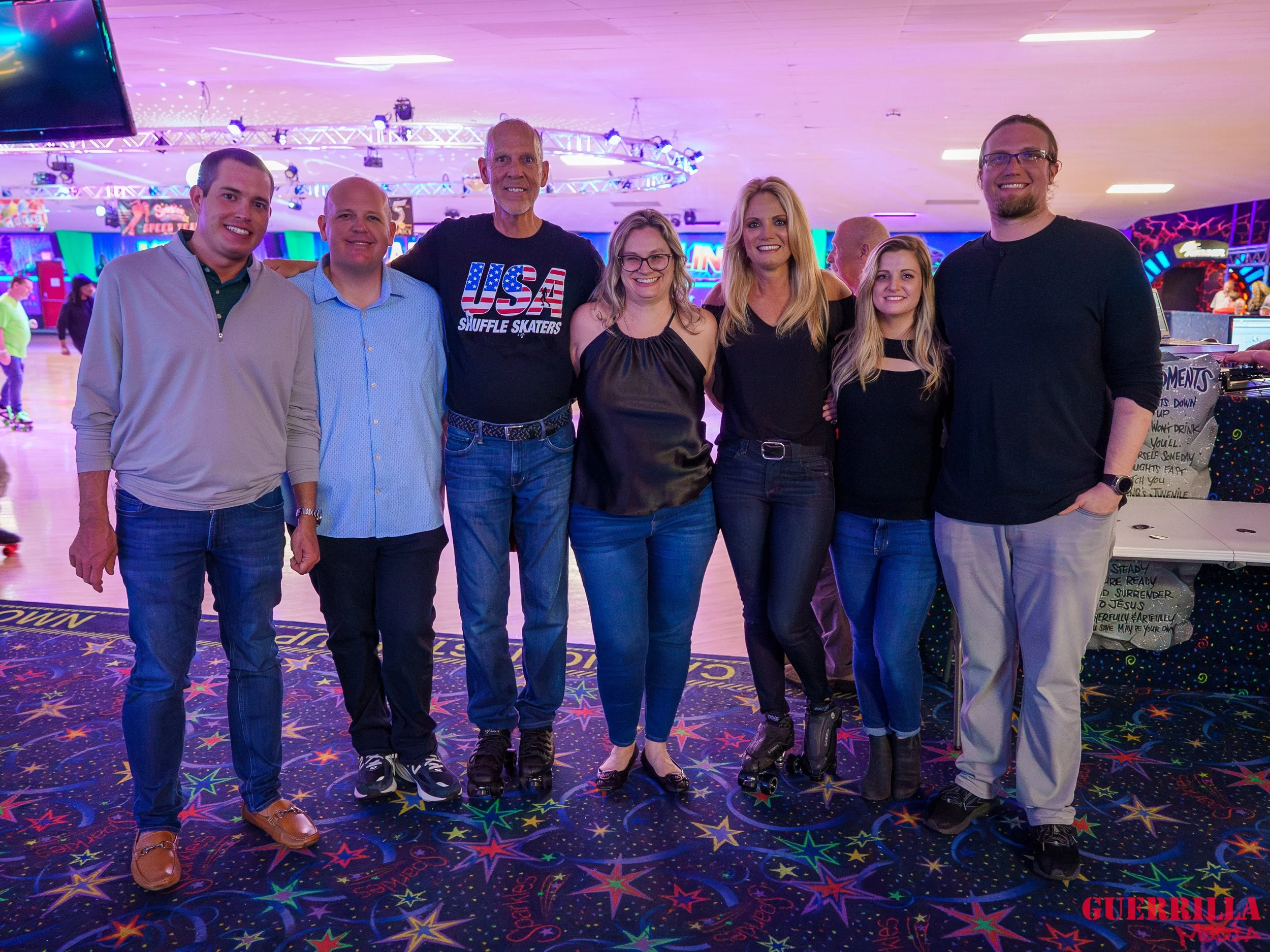 Group of seven people standing together at an indoor roller skating rink, smiling for the camera, with the rink's colorful lights and skating area in the background.