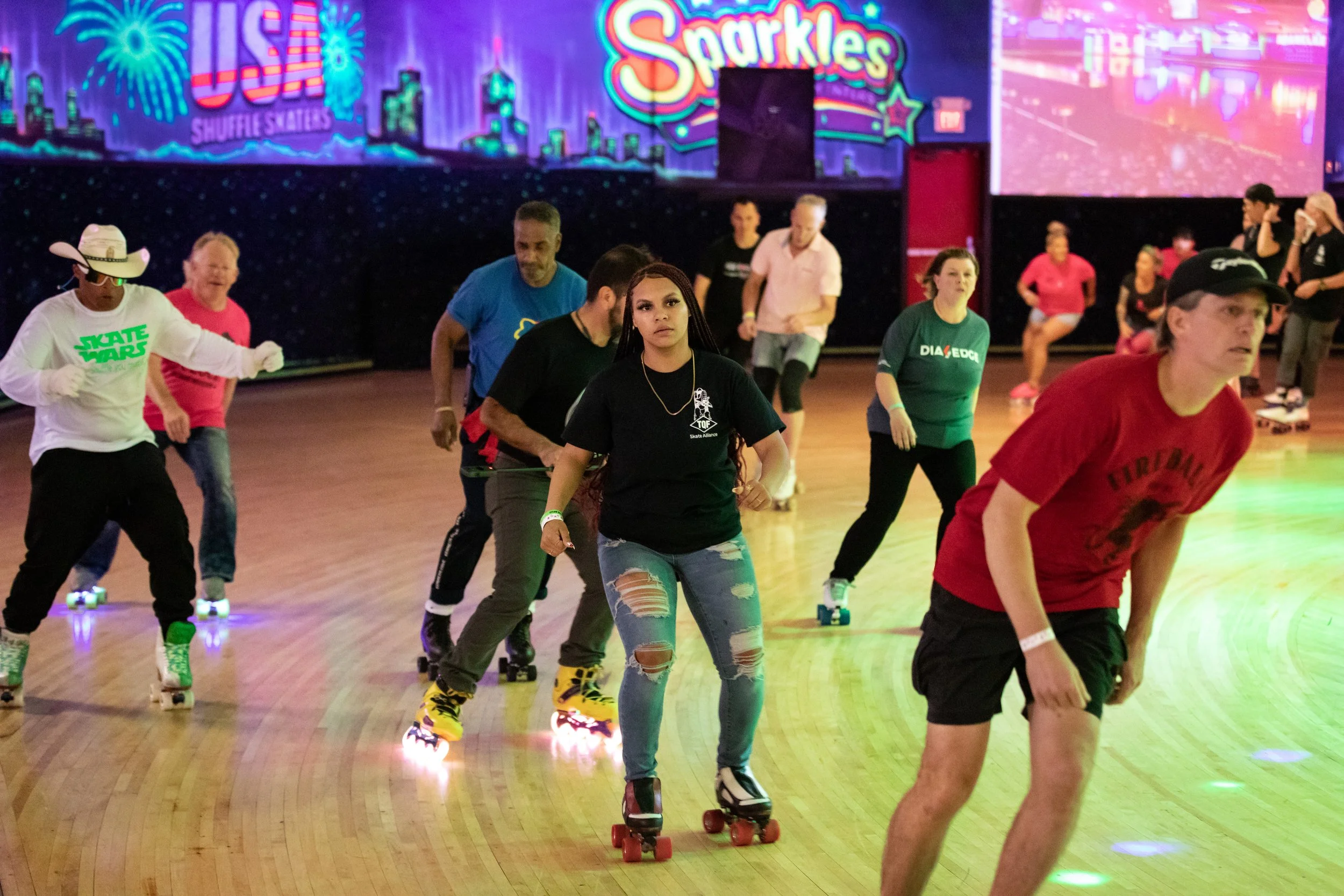 People roller skating in a brightly lit skating rink with colorful neon signs and graphics on the walls, including 'US Shuffle Skaters,' and a large screen displaying lights.