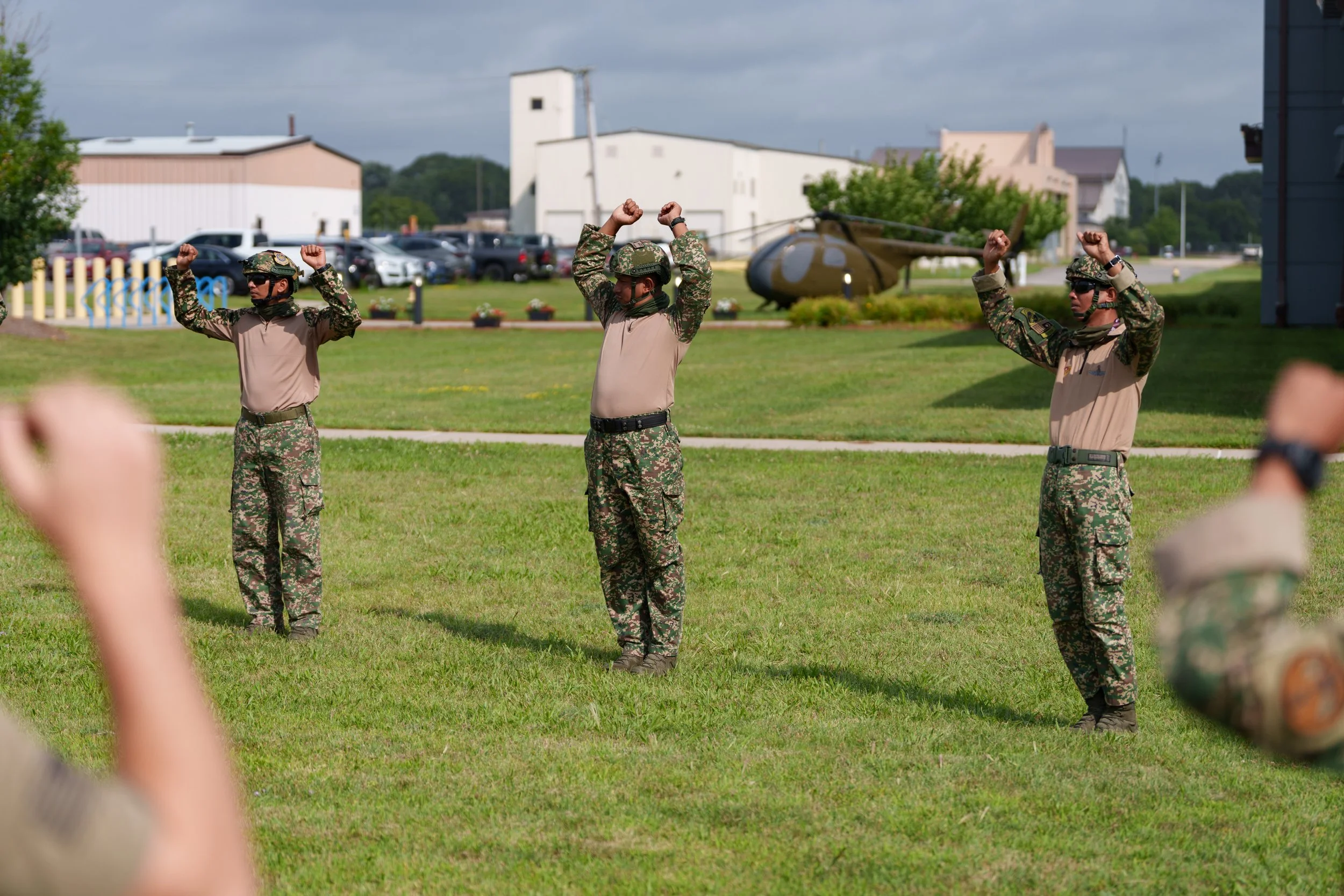 Military personnel in camouflage uniform participating in a training or drill exercise outdoors on a grassy area, with a helicopter and buildings in the background.