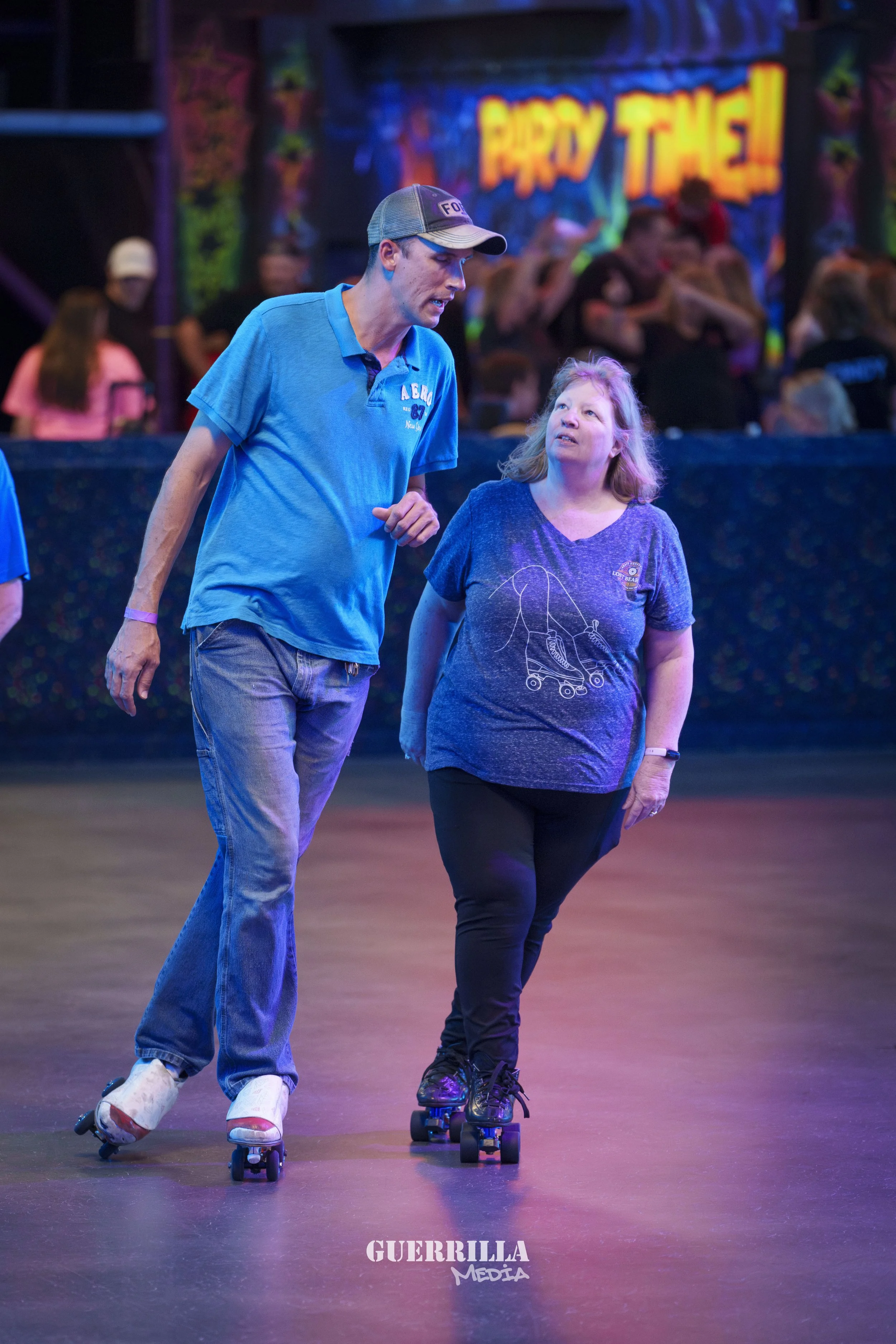 A man and a woman roller skating together at an indoor skating rink with colorful lights. The man is wearing a gray cap and a blue polo shirt, while the woman has long blonde hair and is wearing a purple T-shirt with a roller skate graphic. They are 