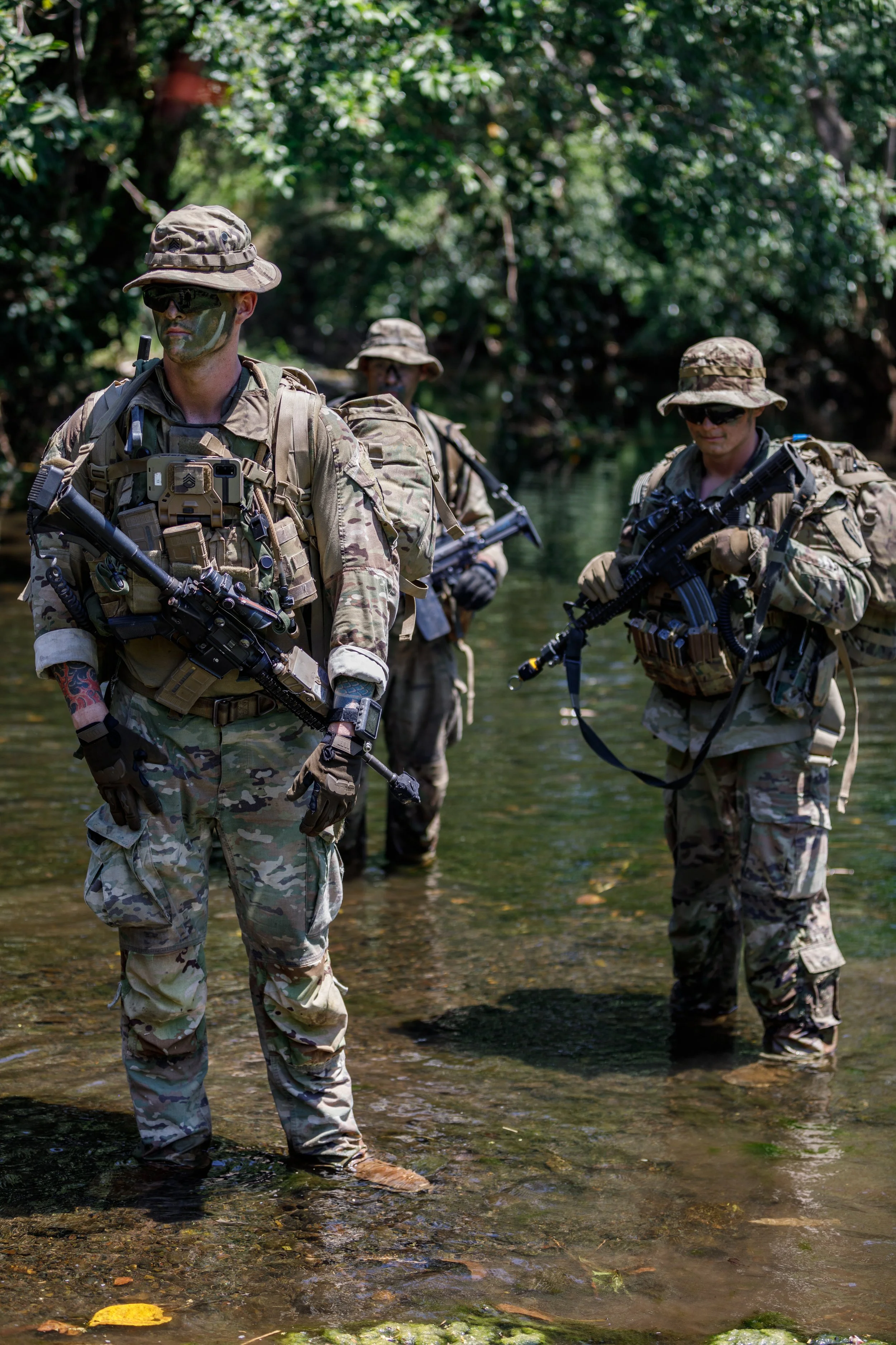 Group of soldiers in camouflage uniforms wading through a shallow river in a forested area.
