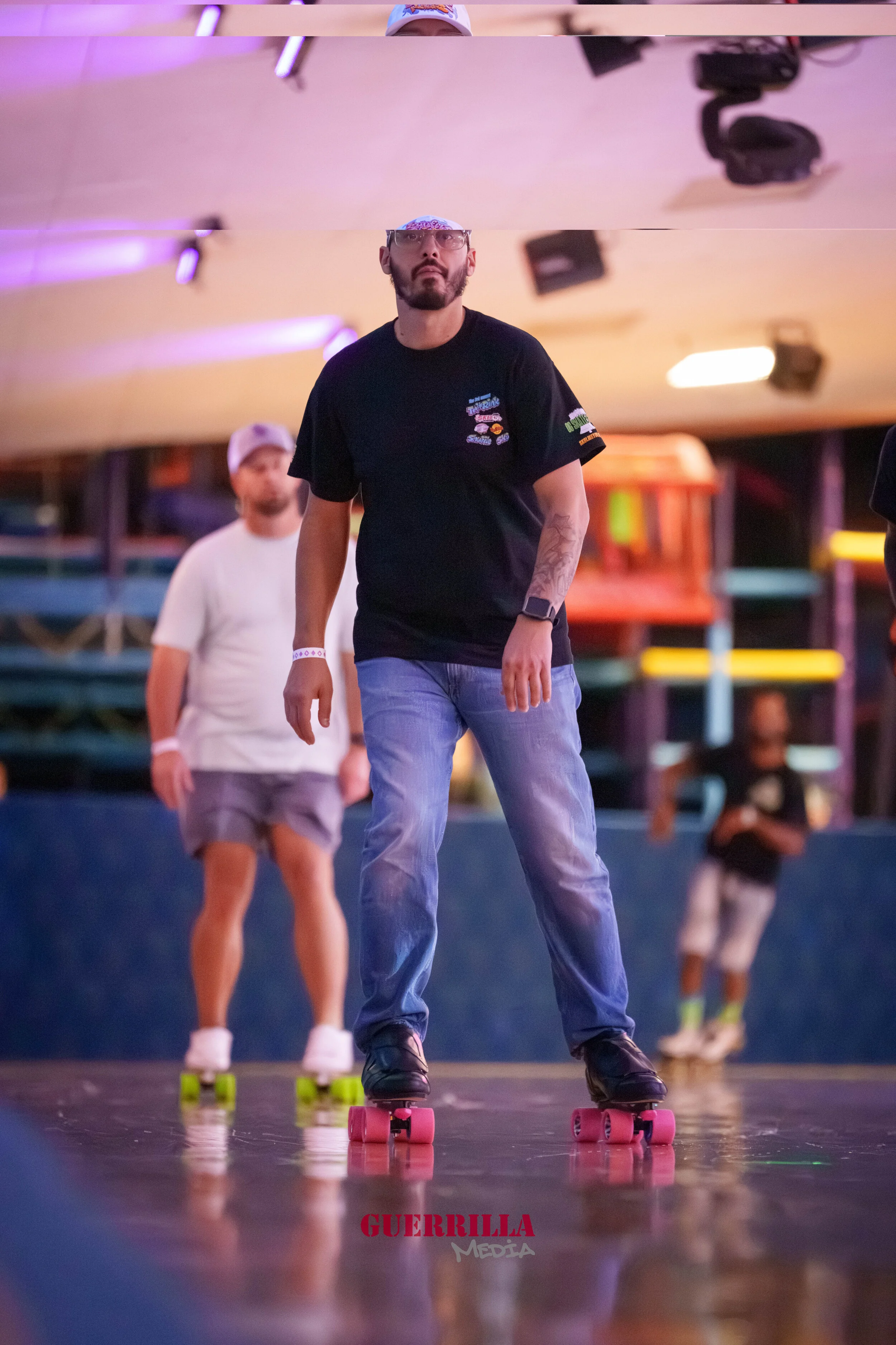 Three people roller skating indoors under colorful lights, with the focus on a man in a black t-shirt and blue jeans in the foreground. 