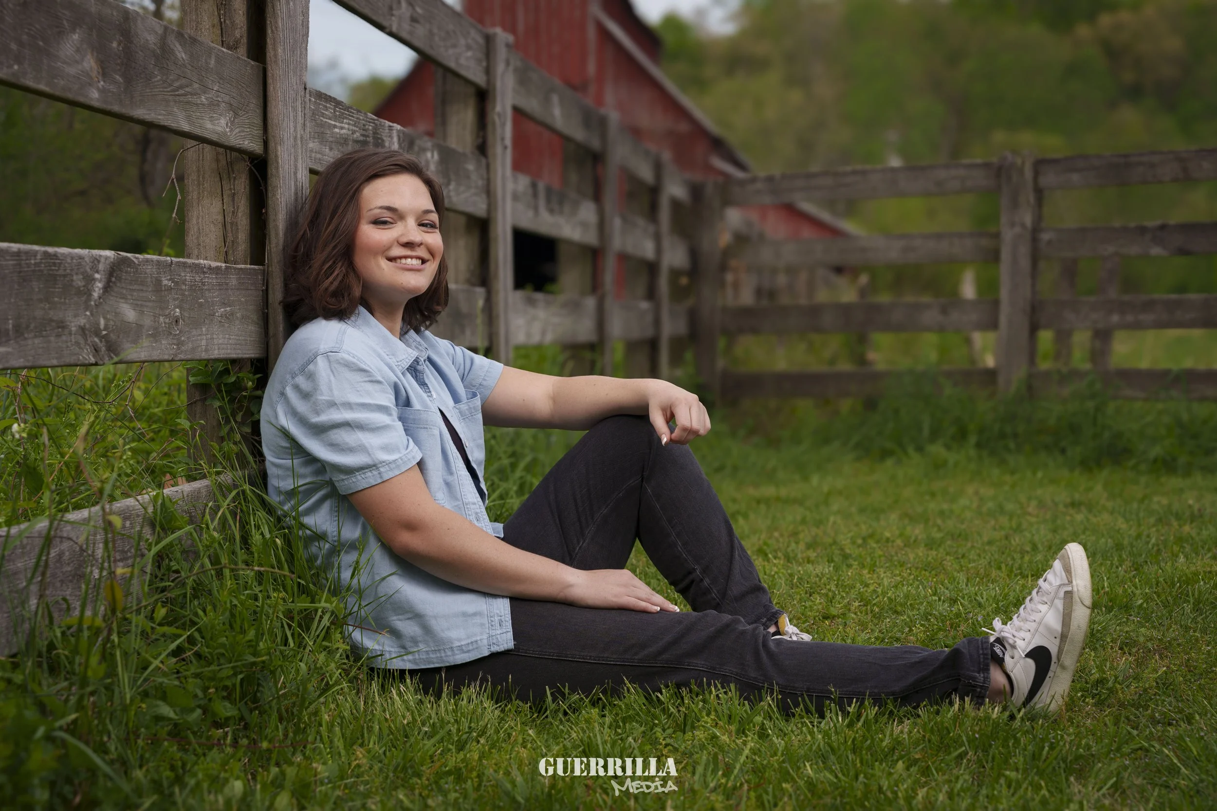 A young woman with short brown hair and a light blue denim shirt sitting on grass, leaning against a wooden fence, smiling at the camera, with a red barn and trees in the background.