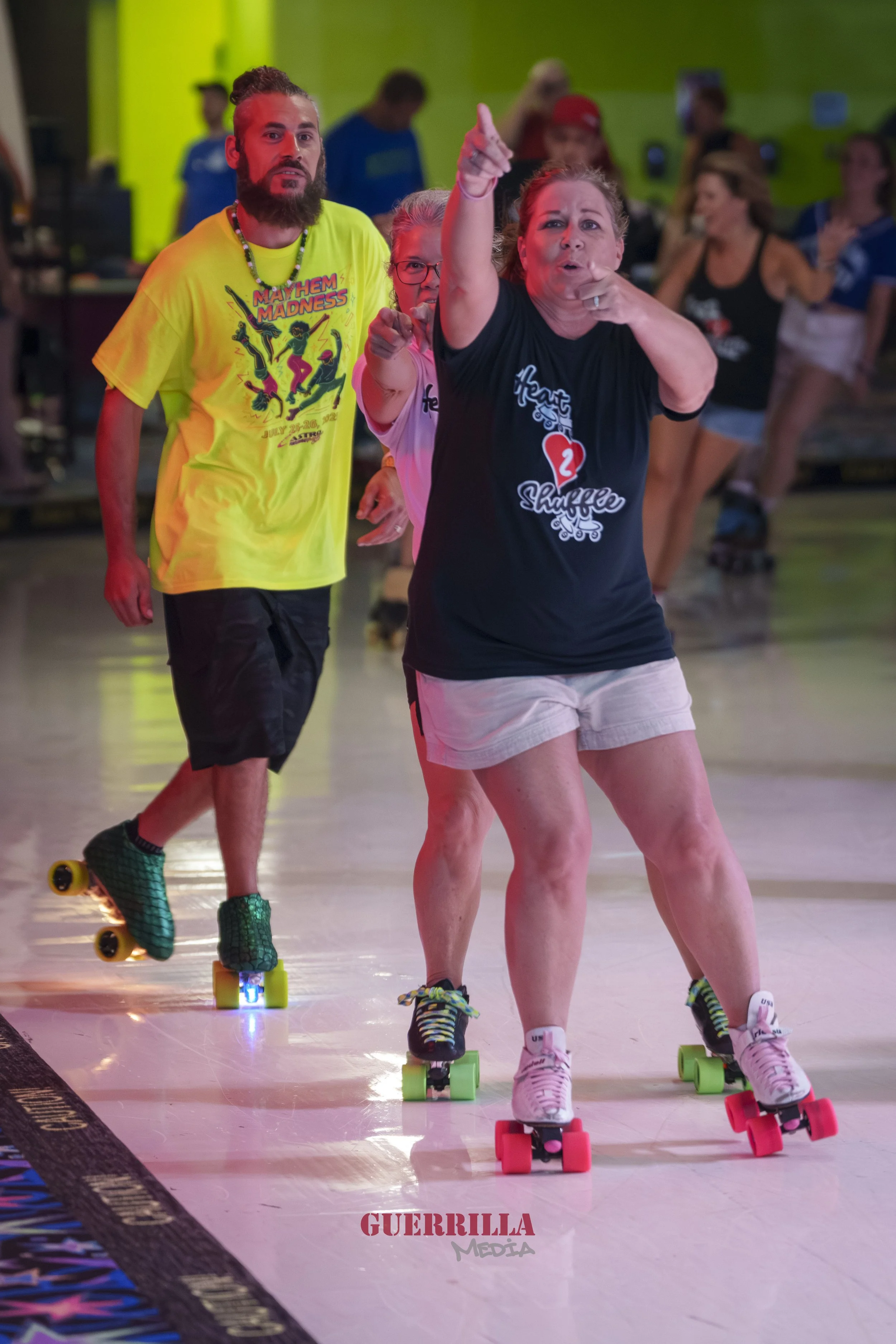 People roller skating indoors, participating in an event called 'Mayhem in Madness,' with a woman in front pointing upward and others following behind, some smiling and cheering.