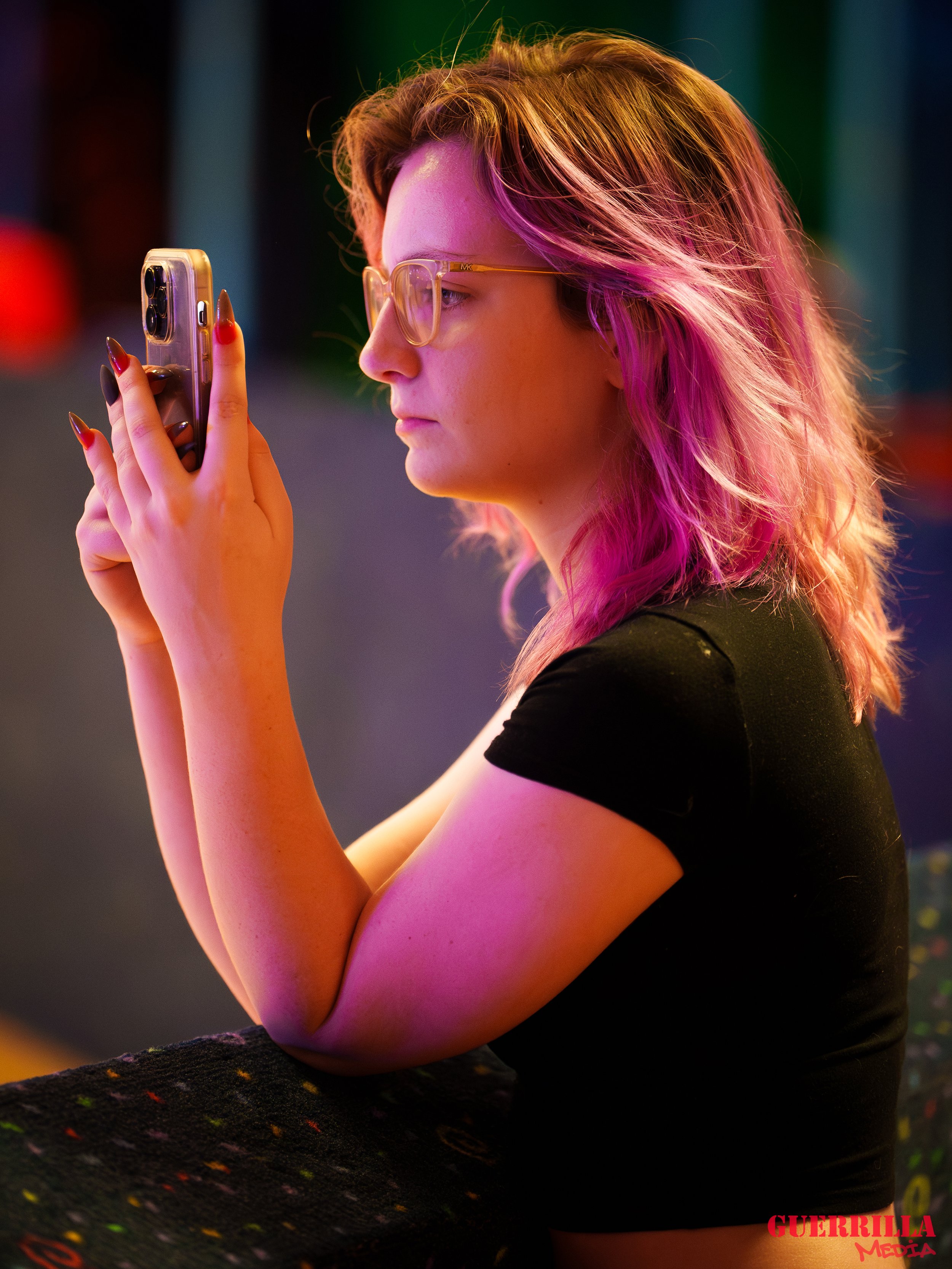 A woman with pink hair and glasses looking at her phone in a dimly lit environment.