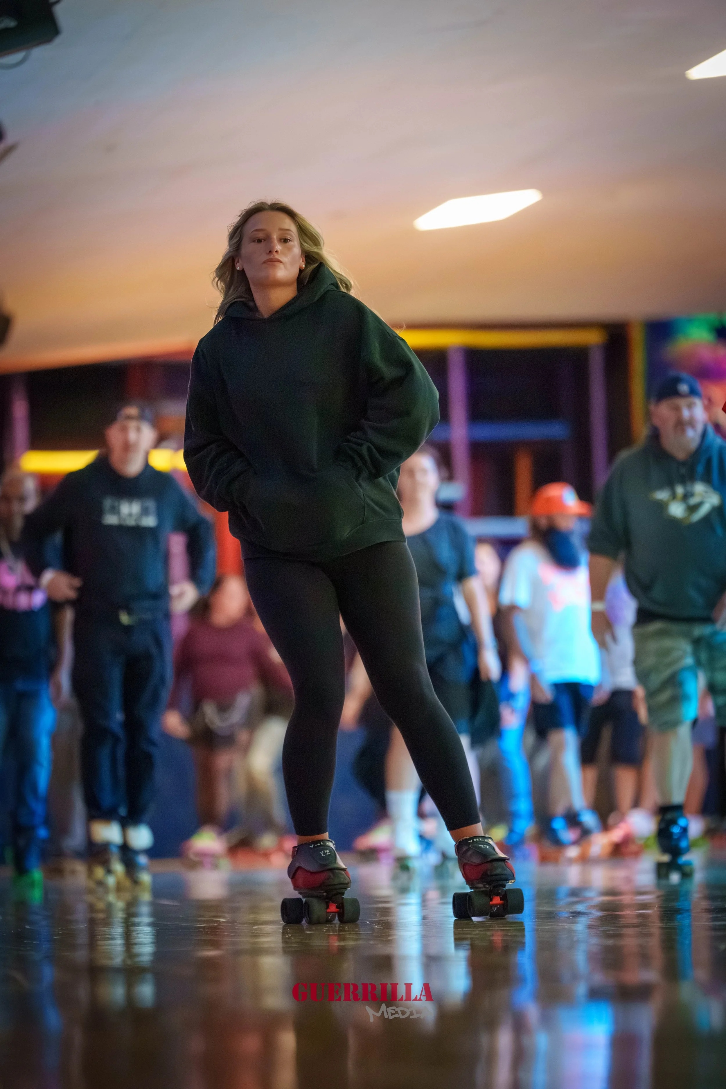 A young woman in black leggings and a dark hoodie roller skating indoors, with other skaters and spectators in the background.