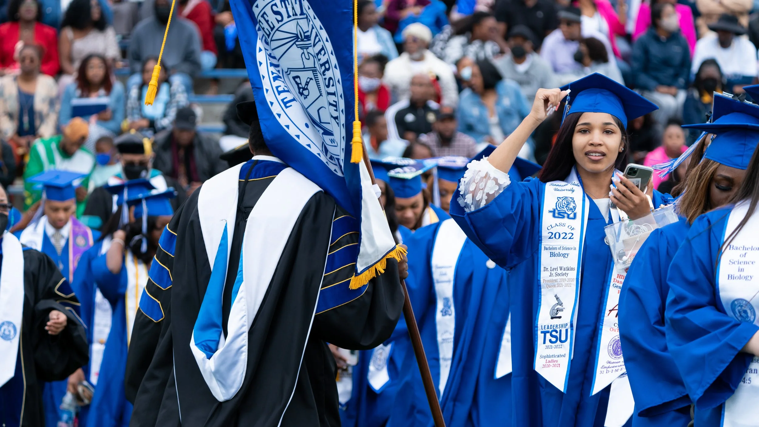 College graduation ceremony with graduates in blue caps and gowns, some wearing ribbons and holding a flag, gathering in an outdoor venue with spectators in the background.