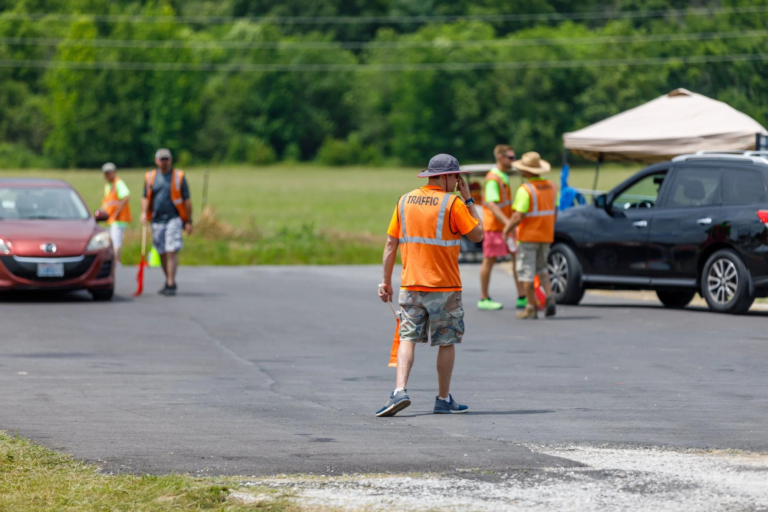Traffic control workers direct traffic at a roadside checkpoint. One worker in the foreground is wearing an orange vest labeled 'TRAFFIC' and talking on a phone, with others in the background near parked cars and a tent, on a rural area with green tr