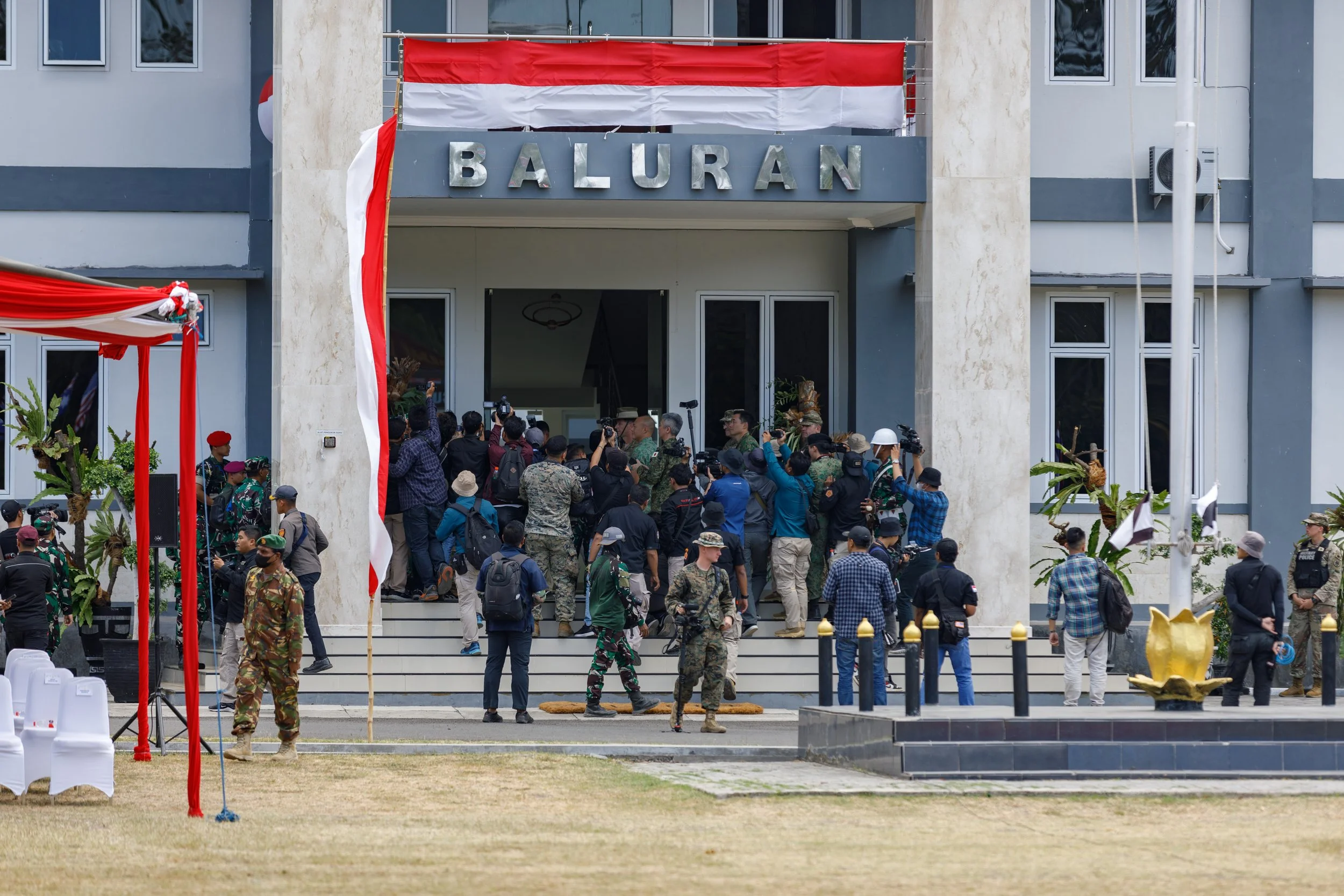 A crowd of people, including military personnel and civilians, gathered outside the entrance of a building labeled "BALURAN," decorated with Indonesian flags and traditional decorations.