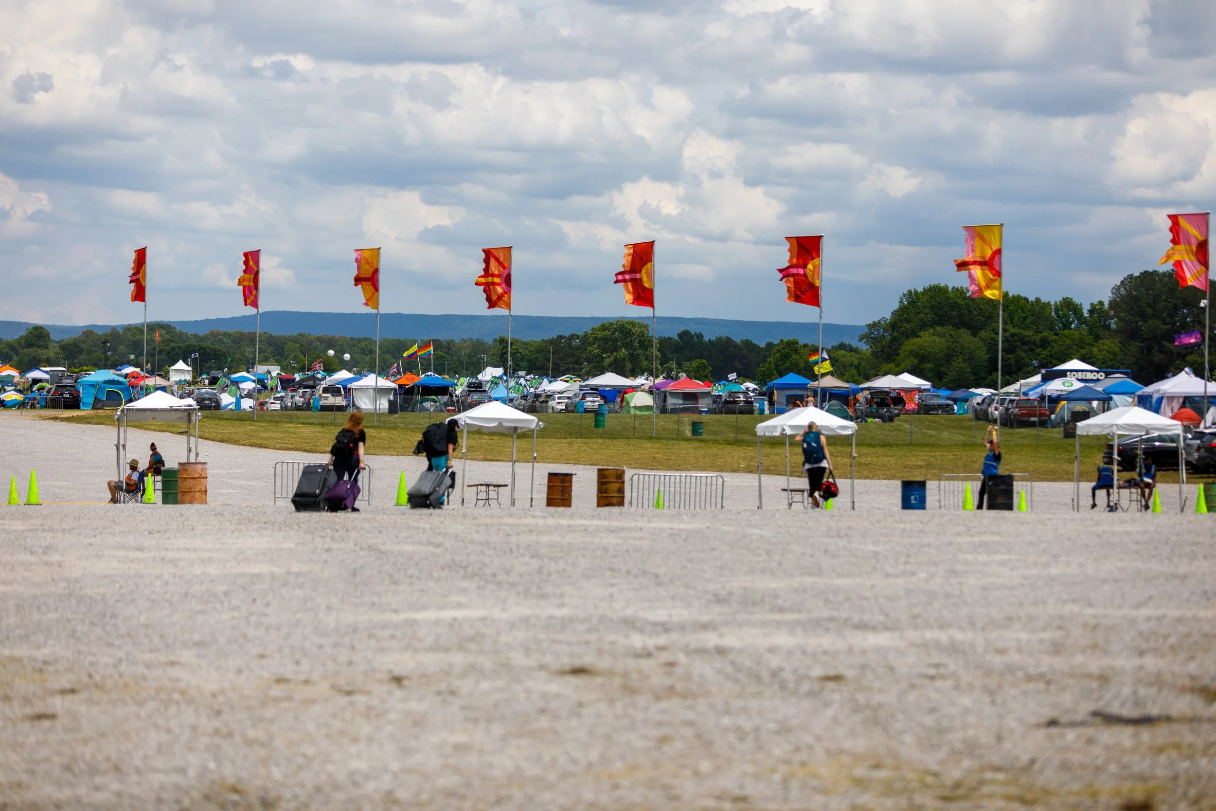 An outdoor event area with tents and flags, some people sitting and walking with luggage, under a partly cloudy sky.