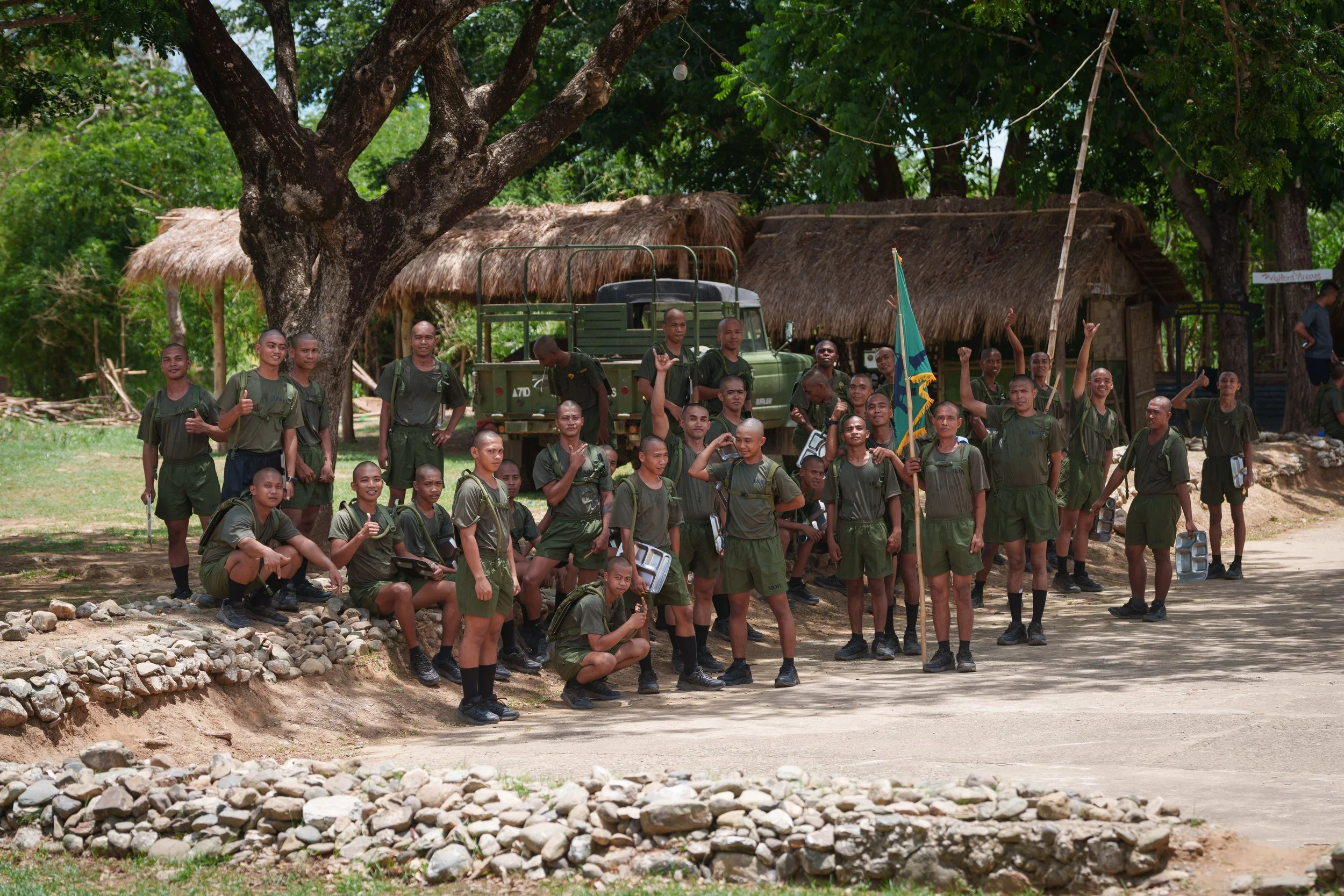 A group of young soldiers in uniform standing outdoors under a large tree, some holding water containers and a flag, with rustic buildings and greenery in the background.
