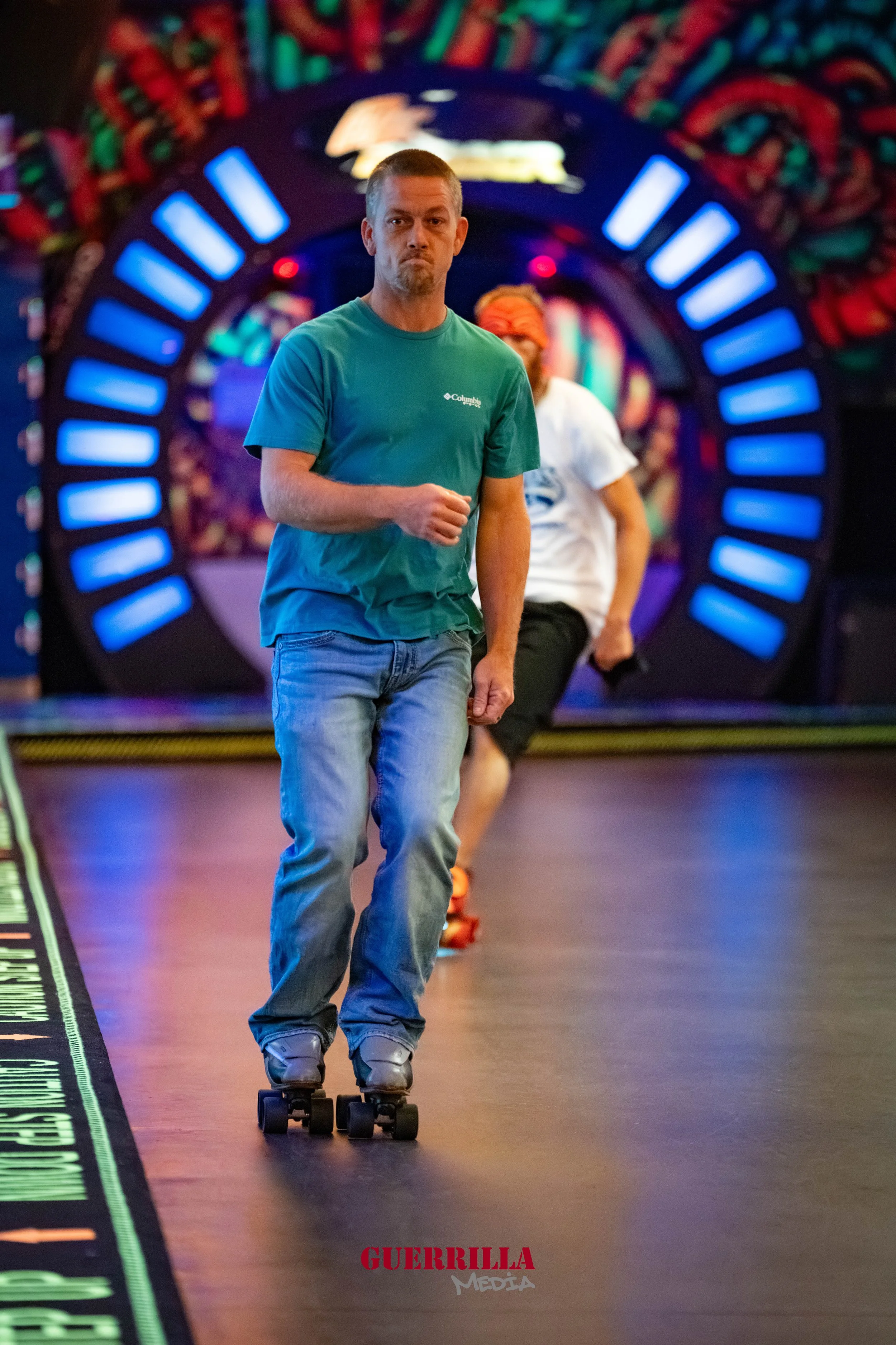 Two people roller skating in a colorful indoor skating rink, with neon lights and vibrant wall art in the background. The person in the foreground is wearing a teal t-shirt and jeans, looking forward with a serious expression.